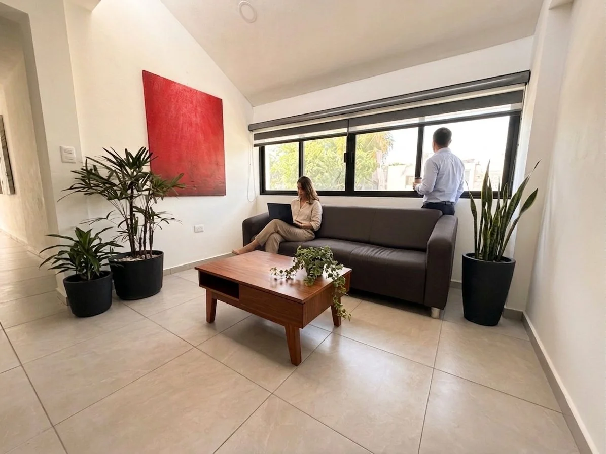 Sala de estar con dos personas: una mujer sentada en un sofá con laptop y un hombre de pie con una taza frente a una ventana grande.