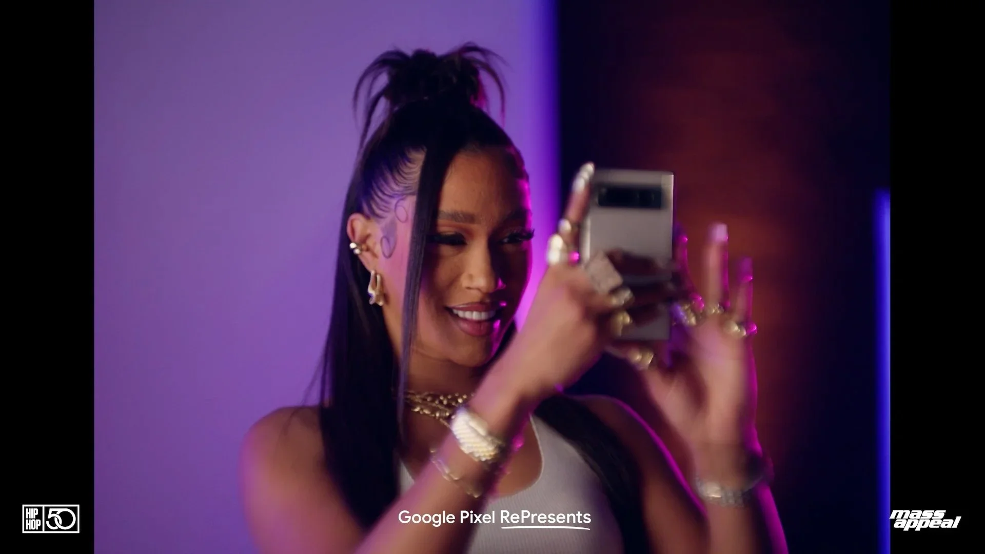 A woman with dark hair styled in a high ponytail and decorated with purple and black face tattoos takes a selfie with her smartphone. She is smiling and wearing multiple jewelry pieces, including earrings, rings, and necklaces, against a dark background with purple and pink lighting.