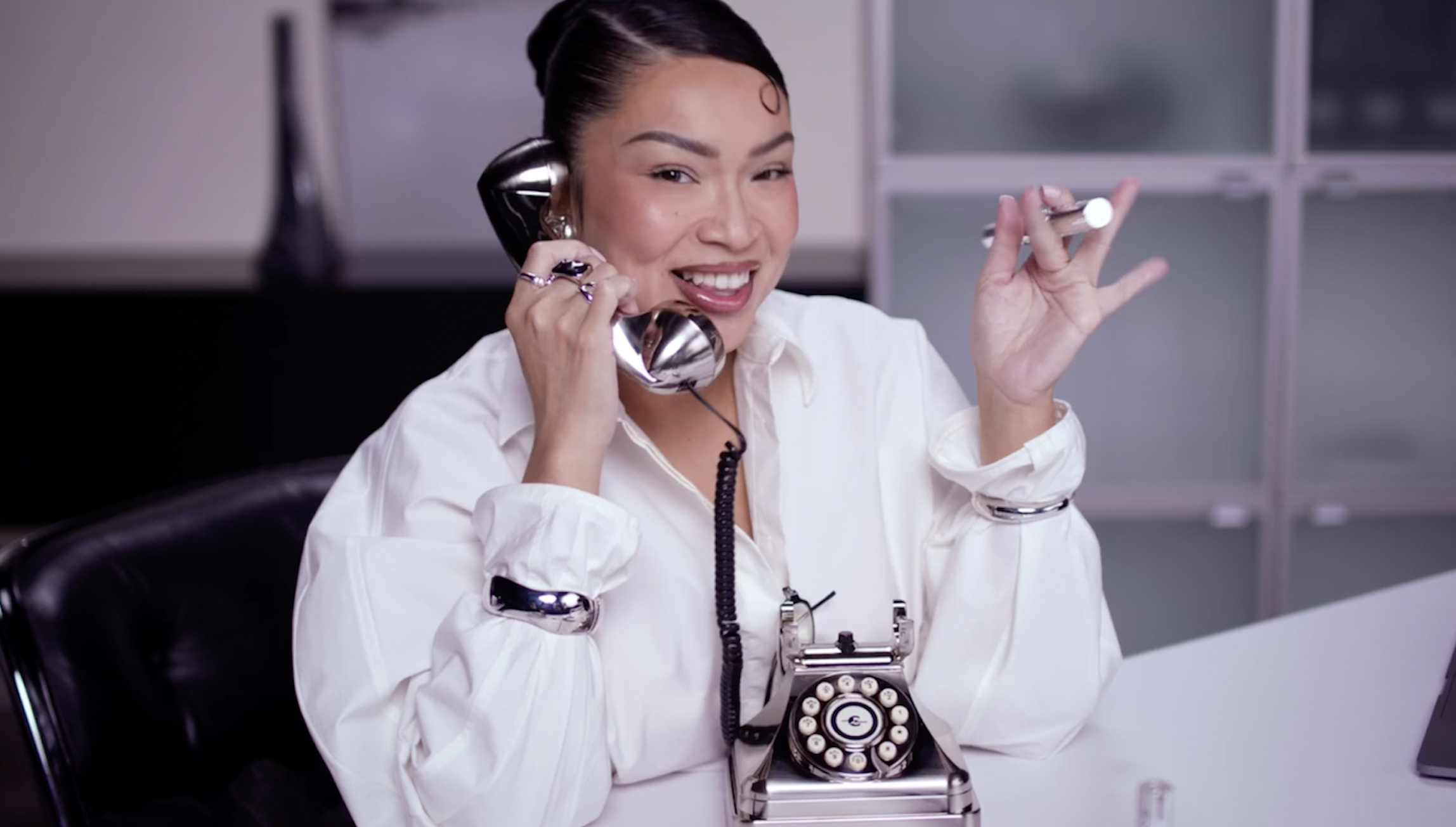Woman with dark hair styled in a bun, smiling and talking on a black rotary phone in an office setting. She is wearing a white blouse and has a vintage camera around her neck.