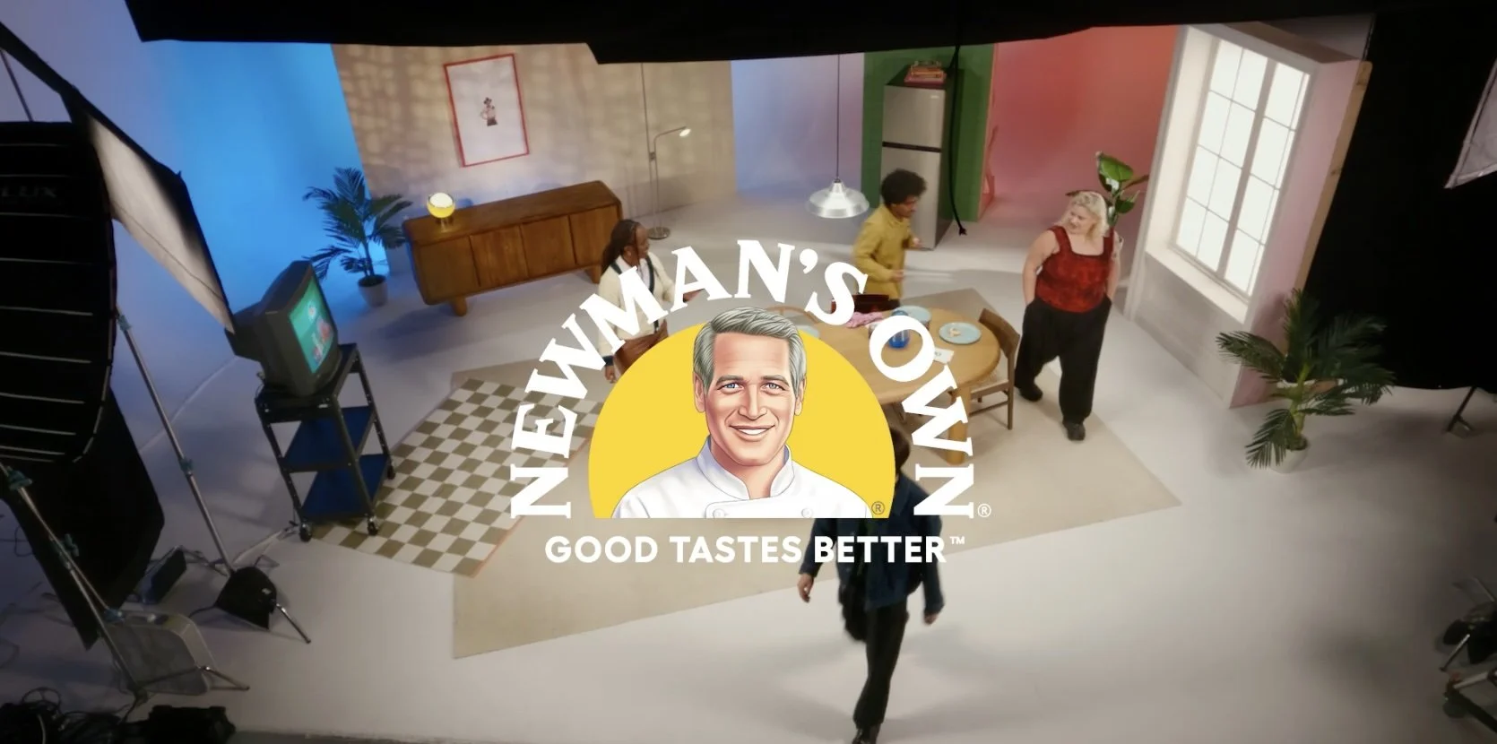 Studio with people setting up a scene for Newman's Own advertisement, including furniture, a TV, and lighting equipment.