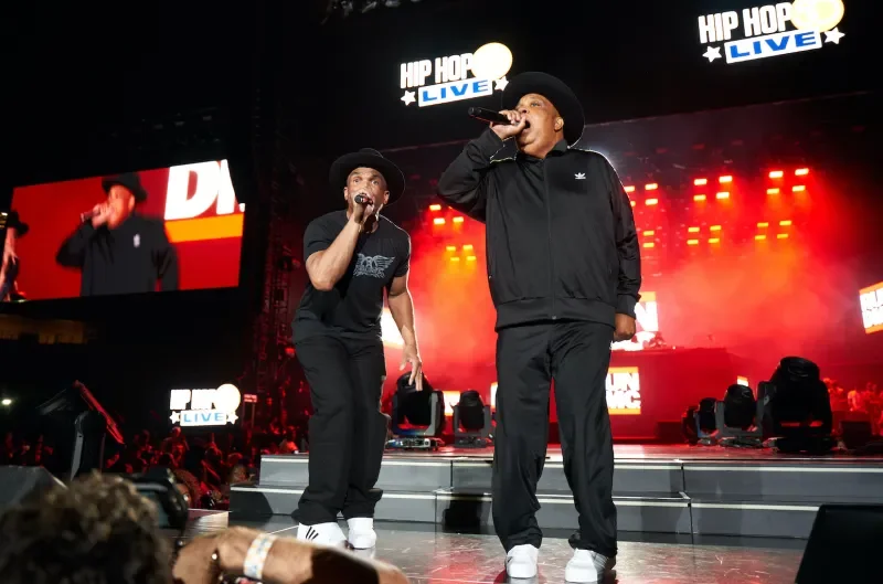 Two male performers on stage singing into microphones at a hip hop concert with red and yellow lights, large screens displaying the event logo, and an audience in front.