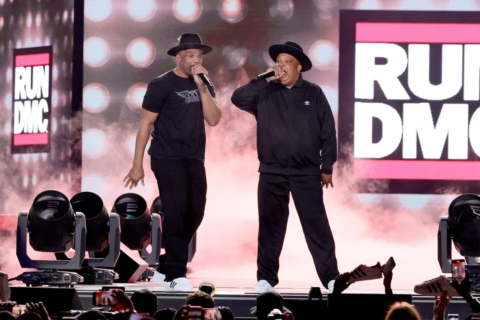 Two men on stage, one wearing a black t-shirt and fedora, and the other in a black jacket and fedora, each holding a microphone, performing in front of a large screen displaying 'RUN DMC' logos with stage lighting and smoke effects.