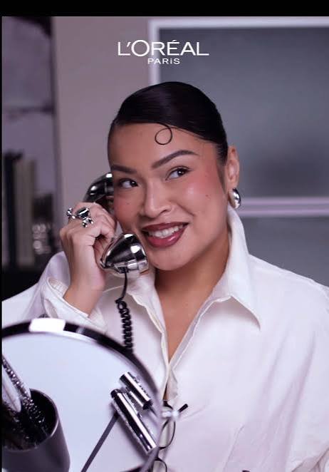 A woman with dark hair styled back and makeup, holding a vintage telephone receiver to her ear, smiling. She is wearing a white shirt and silver jewelry, with a background of a modern interior. The text "L'OREAL PARIS" is at the top of the image.