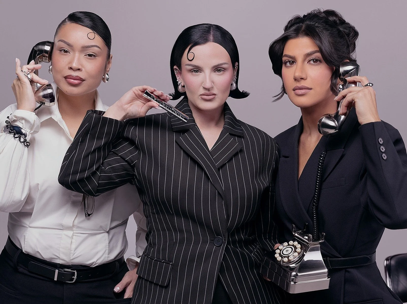 Three women dressed in formal business attire, holding vintage phones to their ears, standing against a plain gray background.
