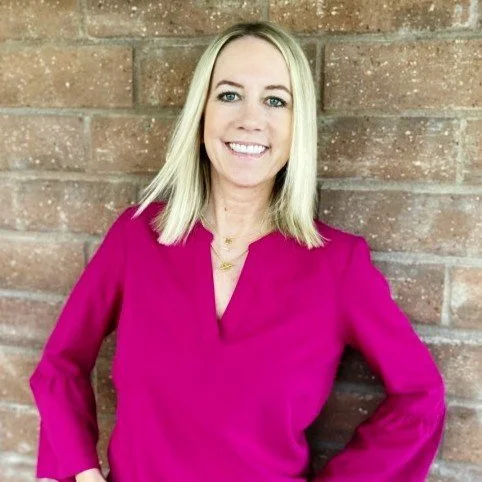 A woman with blonde hair wearing a pink blouse, smiling, standing against a brick wall.