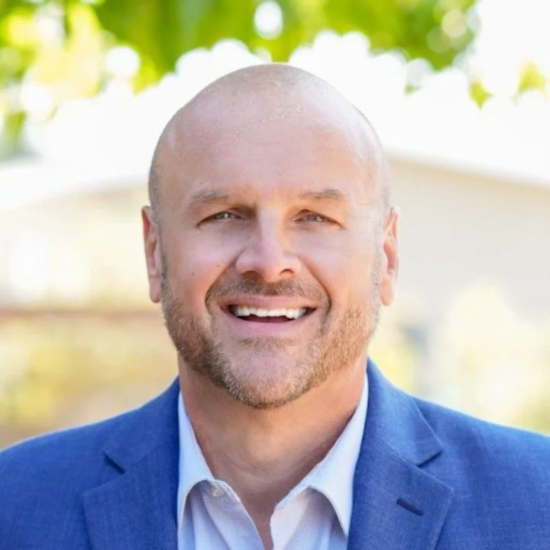 A smiling middle-aged man with a bald head and beard, wearing a blue suit jacket and white shirt, standing outdoors with a blurred background of trees and sunlight.