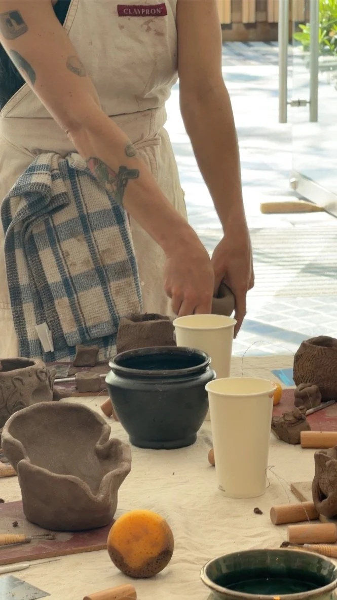 Person working on pottery at a table with bowls, cups, and clay objects, outdoors with sunlight and greenery in the background.