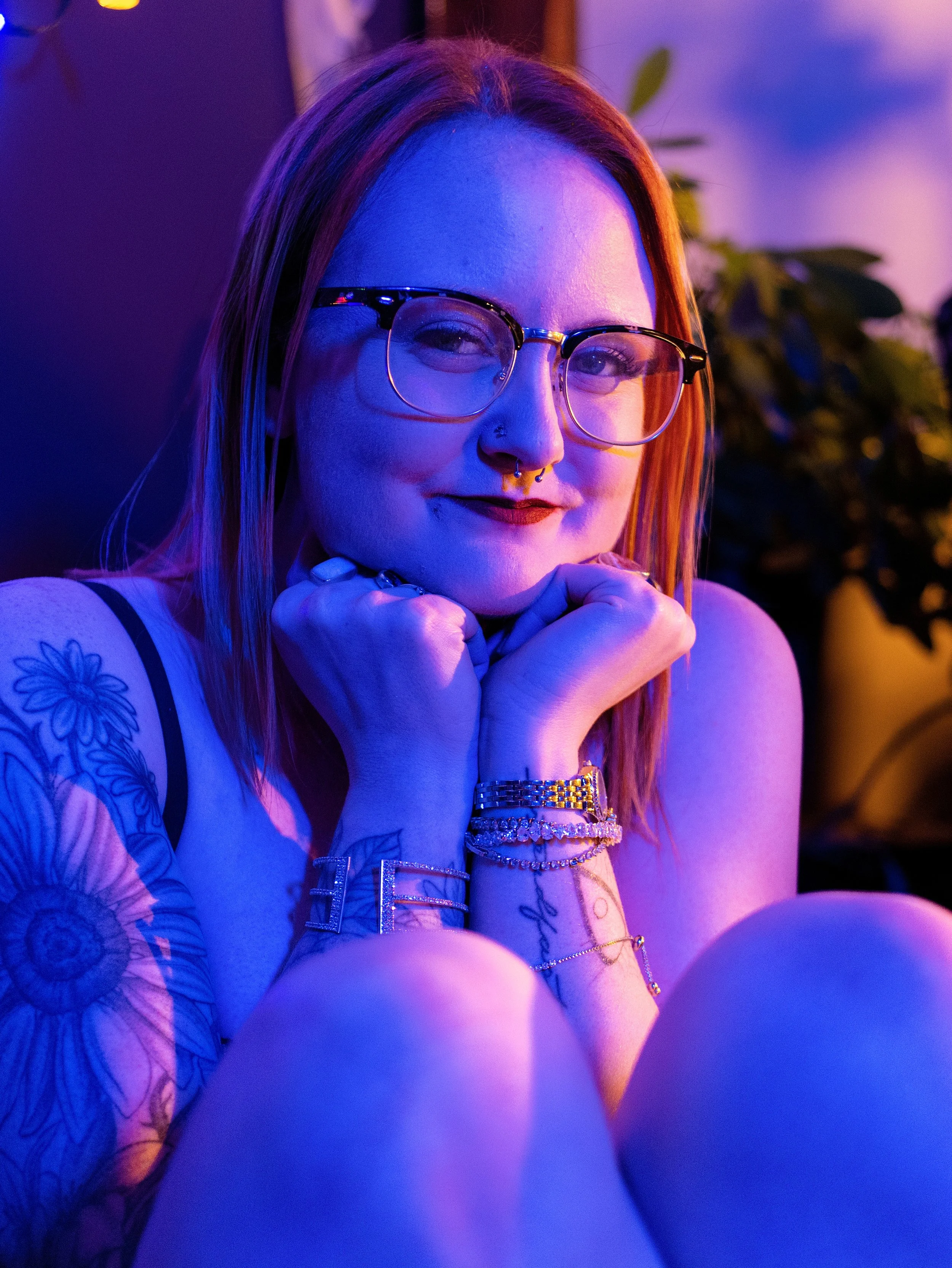 A woman wearing glasses and jewelry, with tattoos on her arms, sitting in a colorful, purple-lit environment.