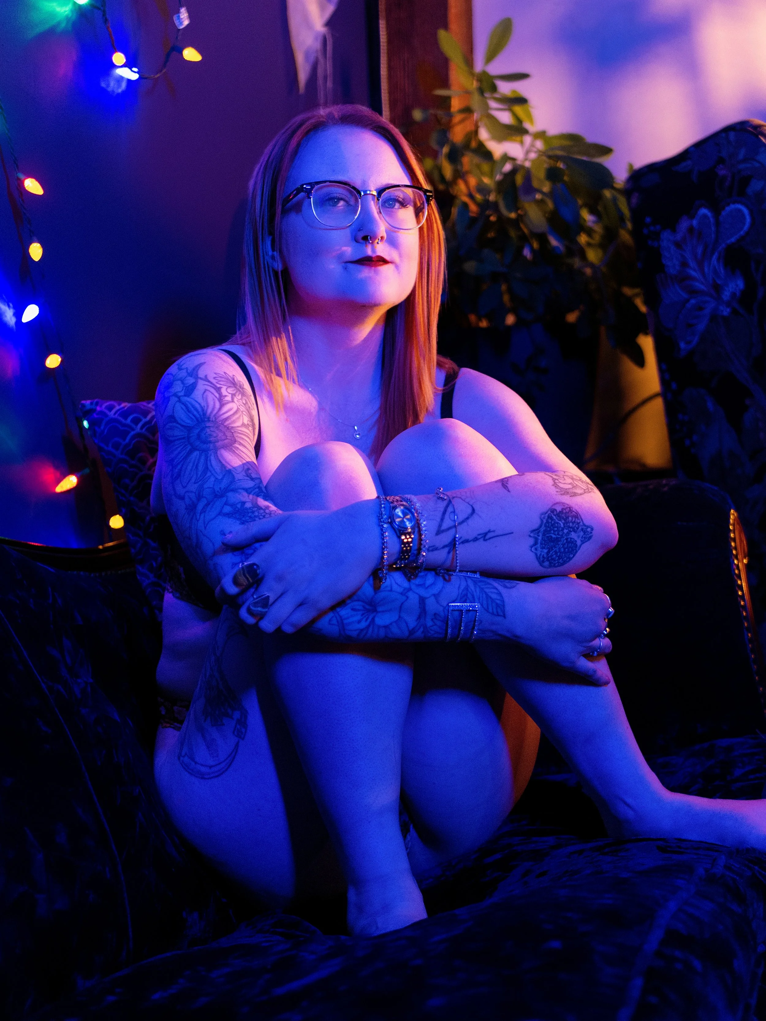 A young woman with red hair, glasses, and tattoos on her arms, sitting with her arms crossed in a room illuminated by colorful, purple-blue lighting and string lights. She has a confident expression and is wearing a tank top, jewelry, and makeup.