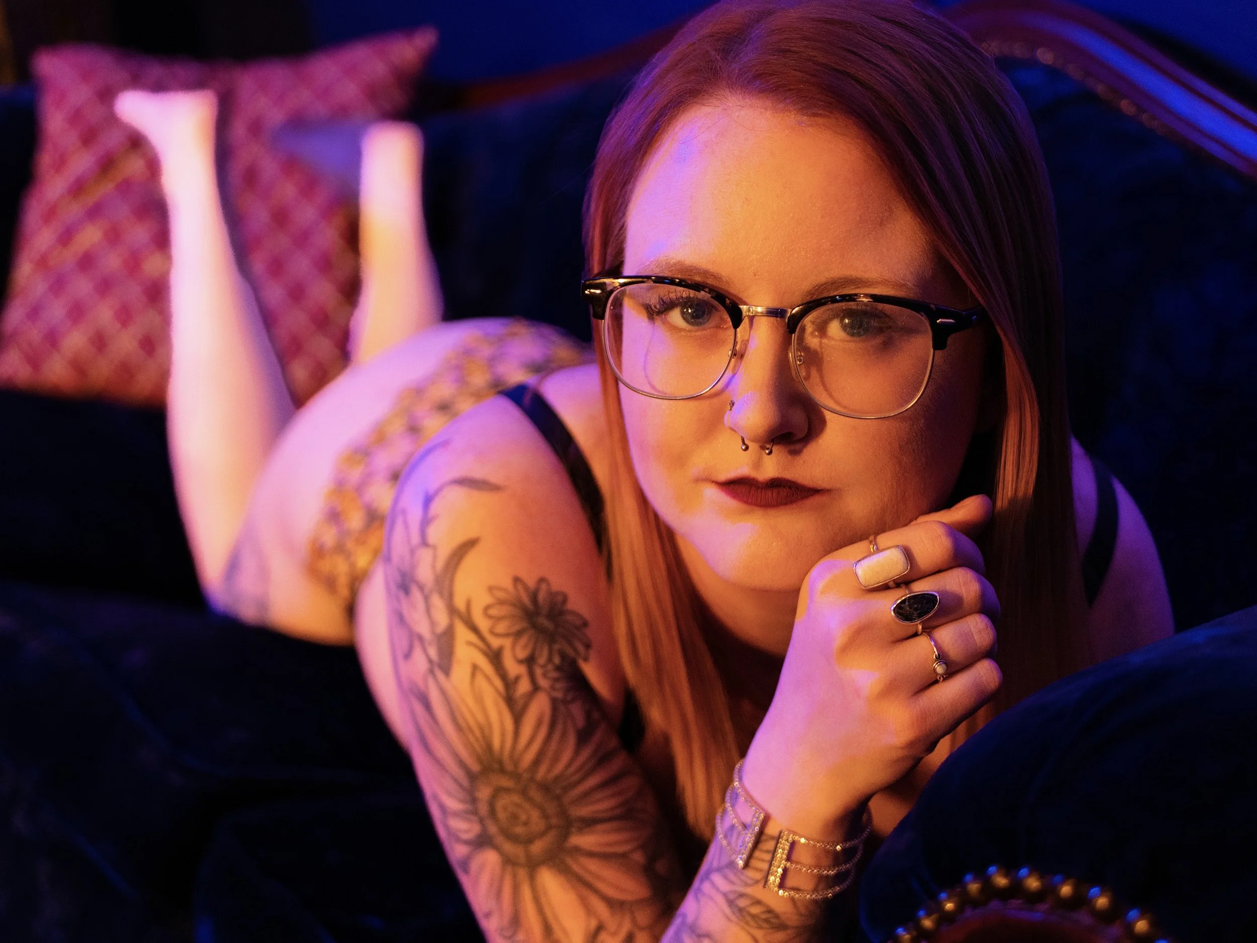 A young woman with glasses and tattoos lying on her stomach on a bed with colorful lighting.