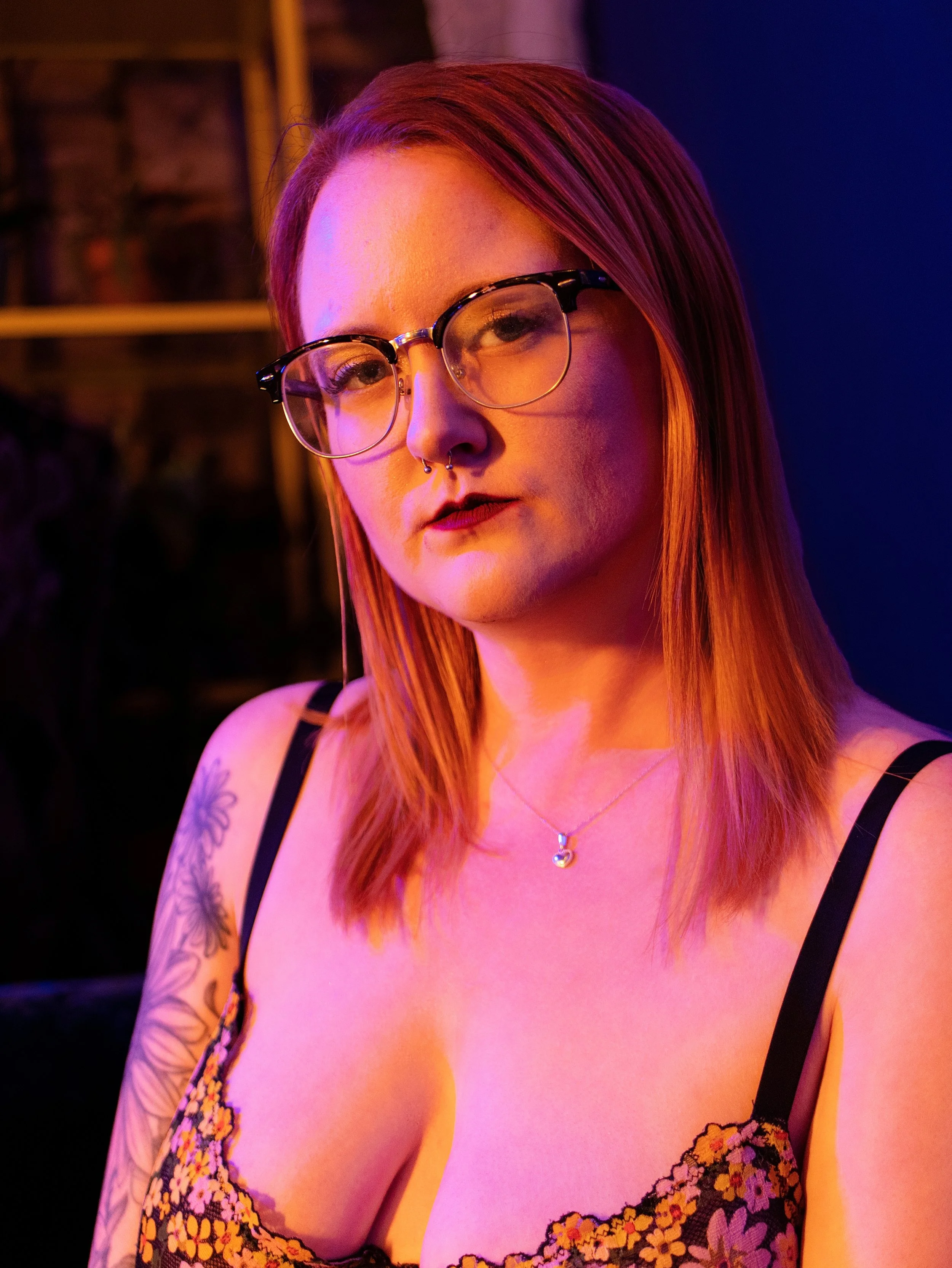 A woman with red hair and glasses, wearing a floral dress and a necklace, posing indoors with colorful lighting.