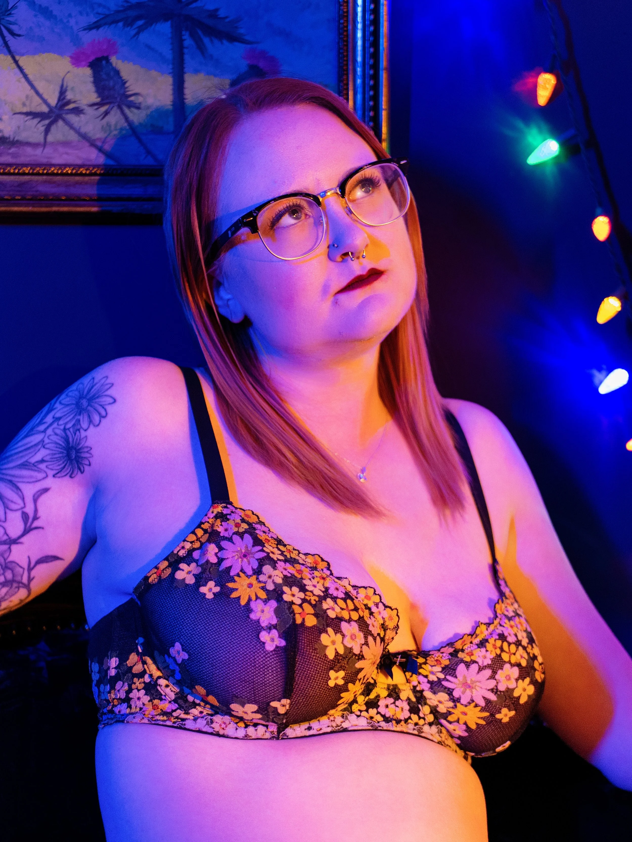 A woman with red hair and glasses looking upward, wearing a floral mesh top, tattoos on her shoulder, and makeup with dark lipstick in a dark room illuminated by colorful string lights and purple-blue lighting.