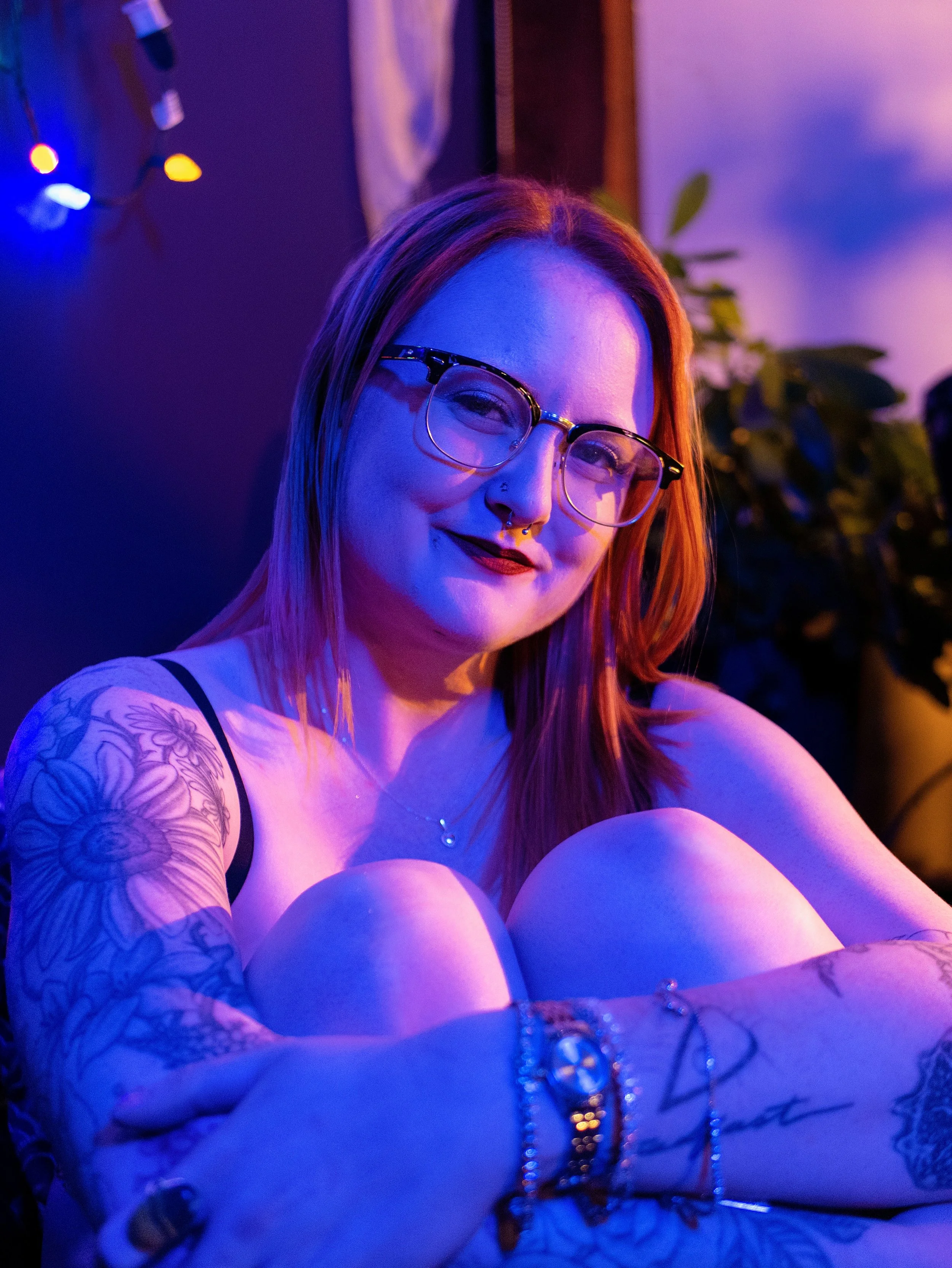 A smiling young woman with glasses and a septum piercing, sitting in a room with blue and purple lighting, tattoos on her arm, and wearing jewelry, with a background of fairy lights and a plant.