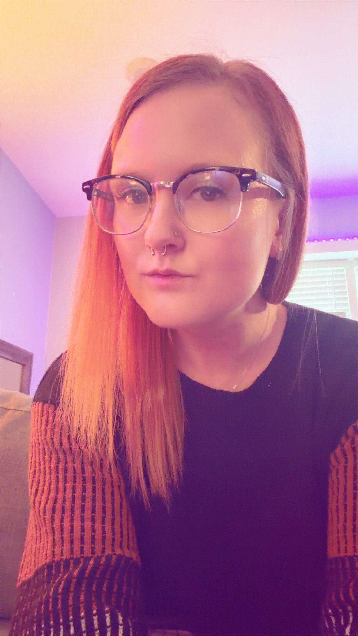 A woman with glasses, long red hair, and pierced nose, sitting indoors with pink and purple lighting in the background.
