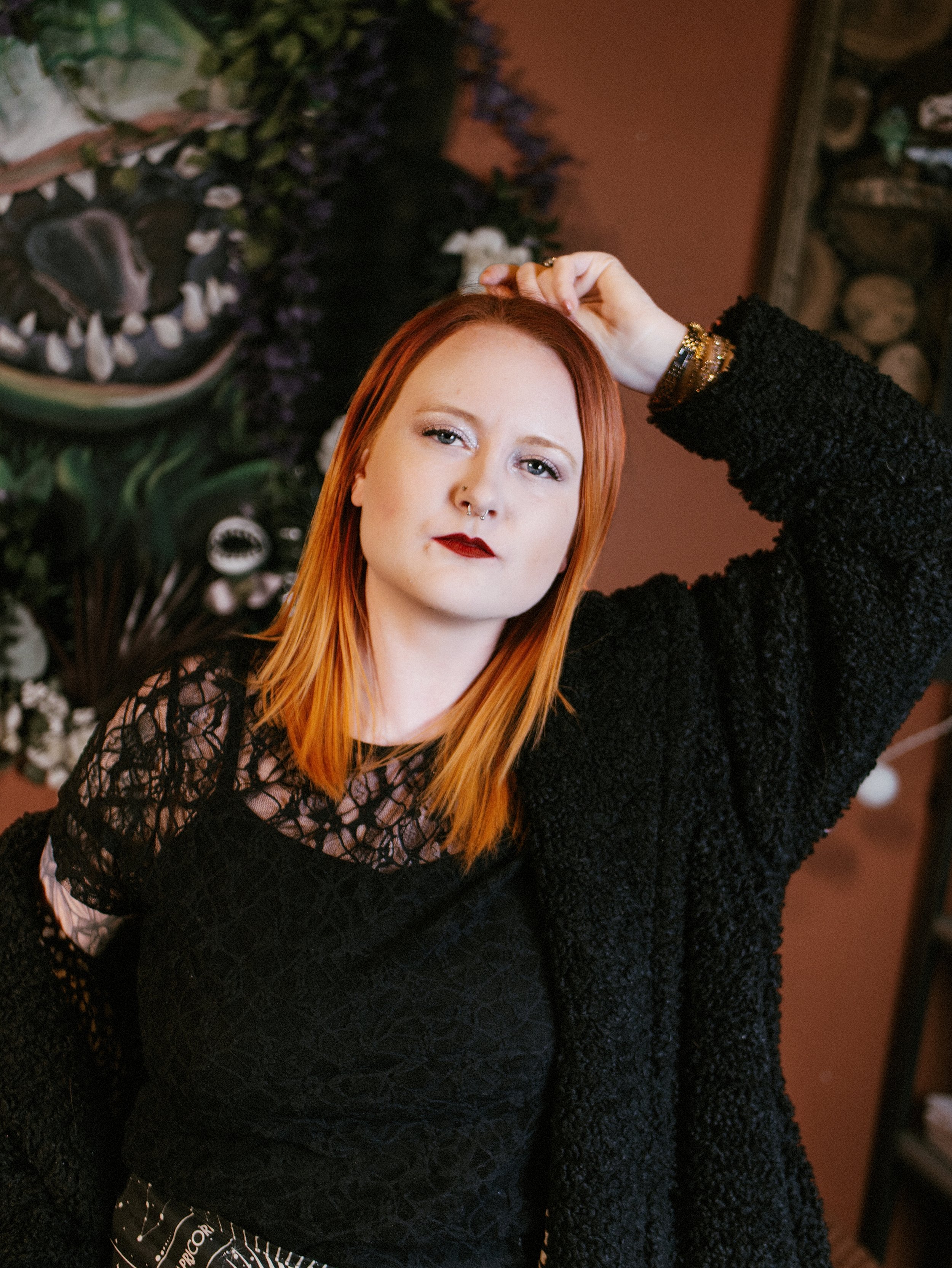 A woman with red hair and fair skin is standing indoors, wearing a black textured top and bracelets, with her hand on her head, looking at the camera.