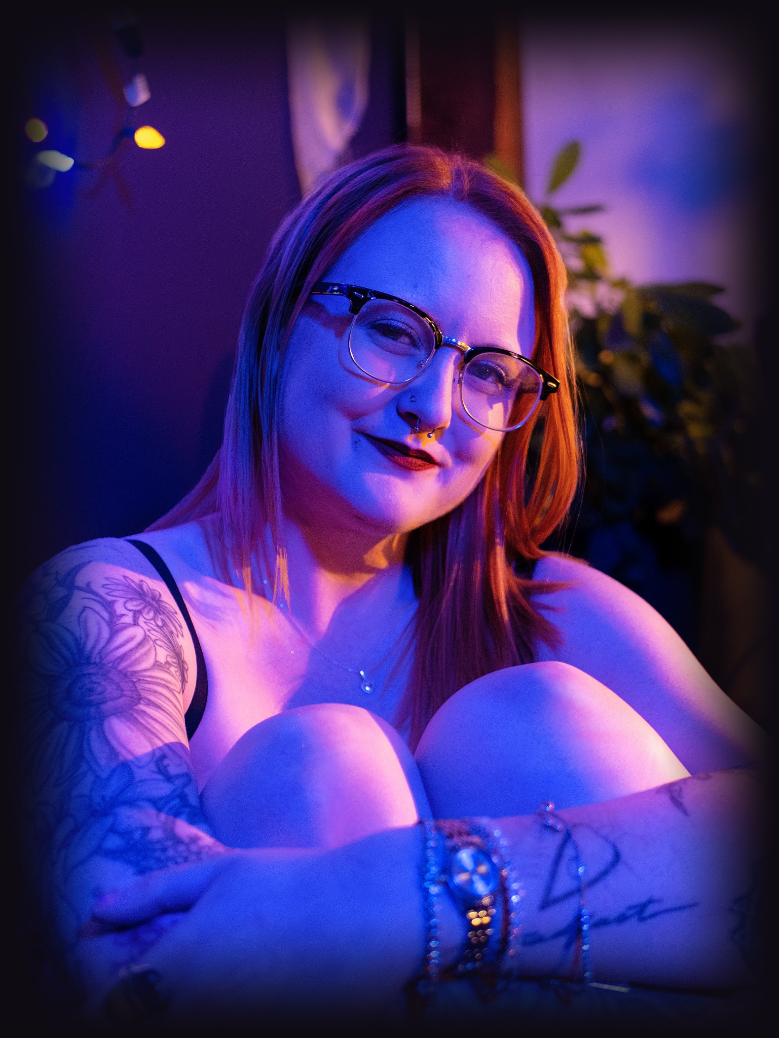 A young woman with red hair, glasses, and a septum piercing, smiling while sitting in a room with blue and purple lighting, tattoos on her arm, wearing a tank top, jewelry, and a bracelet.