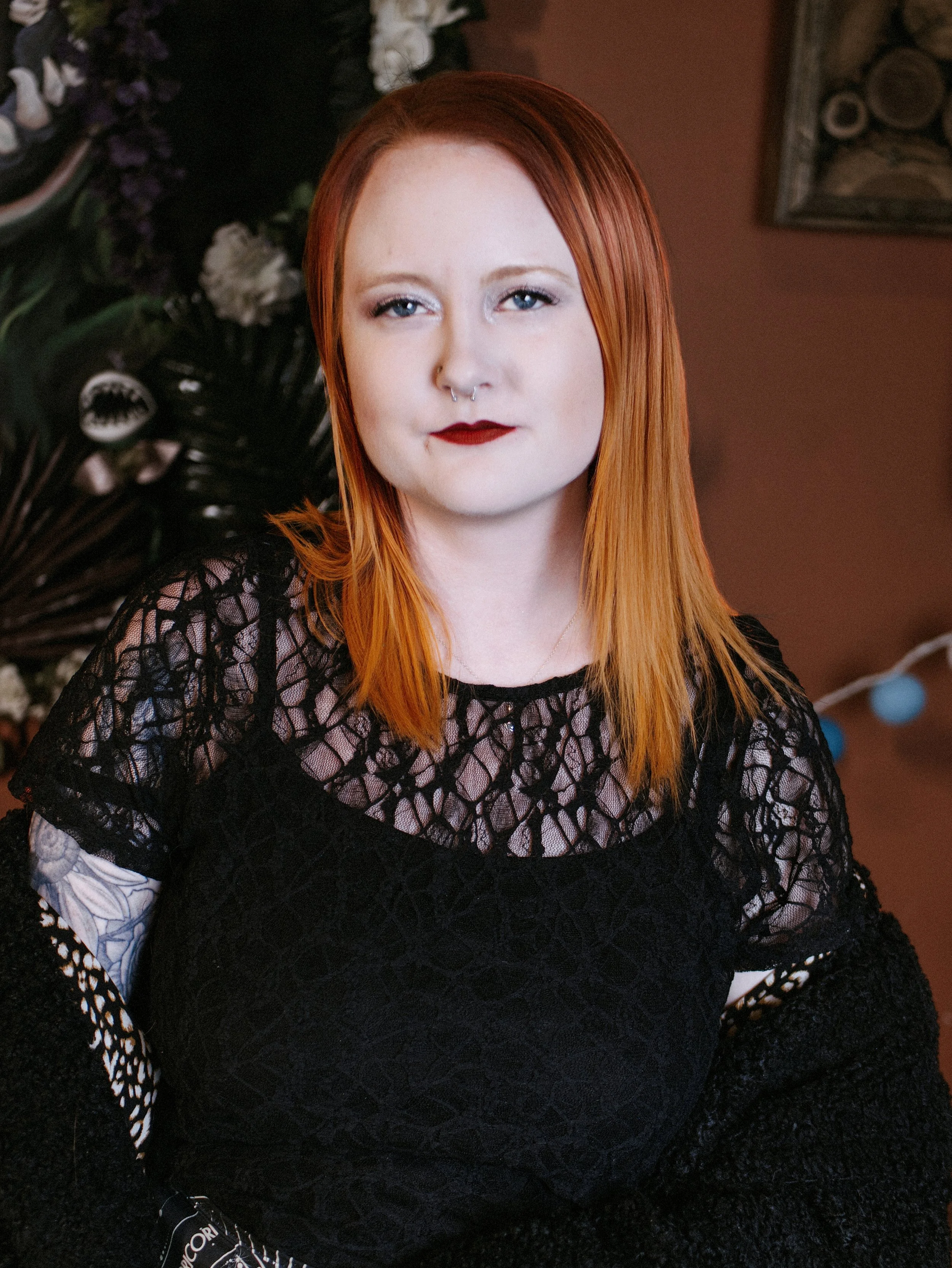 A woman with long red hair and fair skin, wearing red lipstick, a septum piercing, a black lace top, and patterned sleeves, sitting in front of a floral wreath and a brown wall with a framed picture.