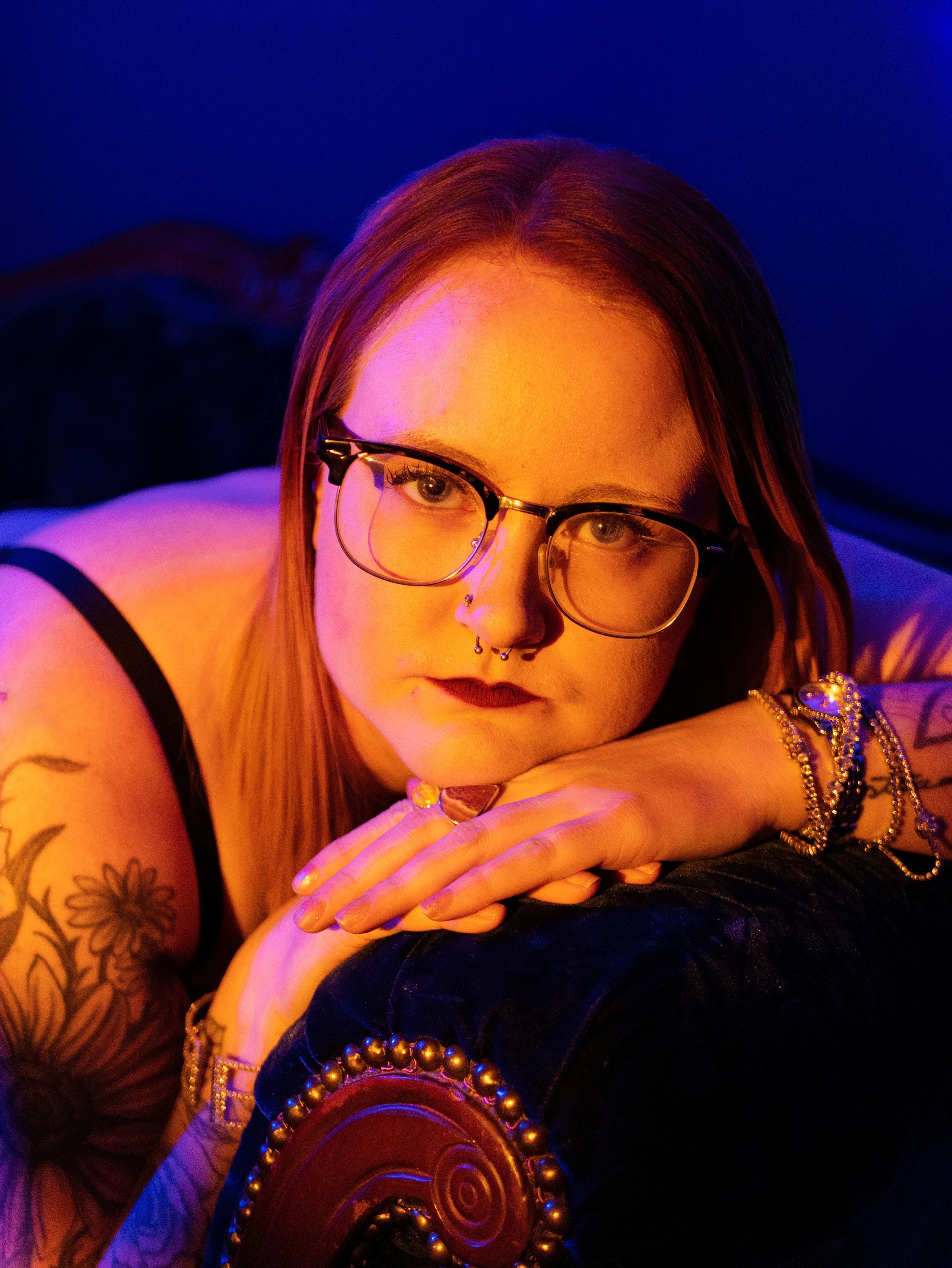 A woman with glasses and long hair, wearing dark lipstick, has her arms crossed on a surface with colorful jewelry and a dark background illuminated by warm lighting.