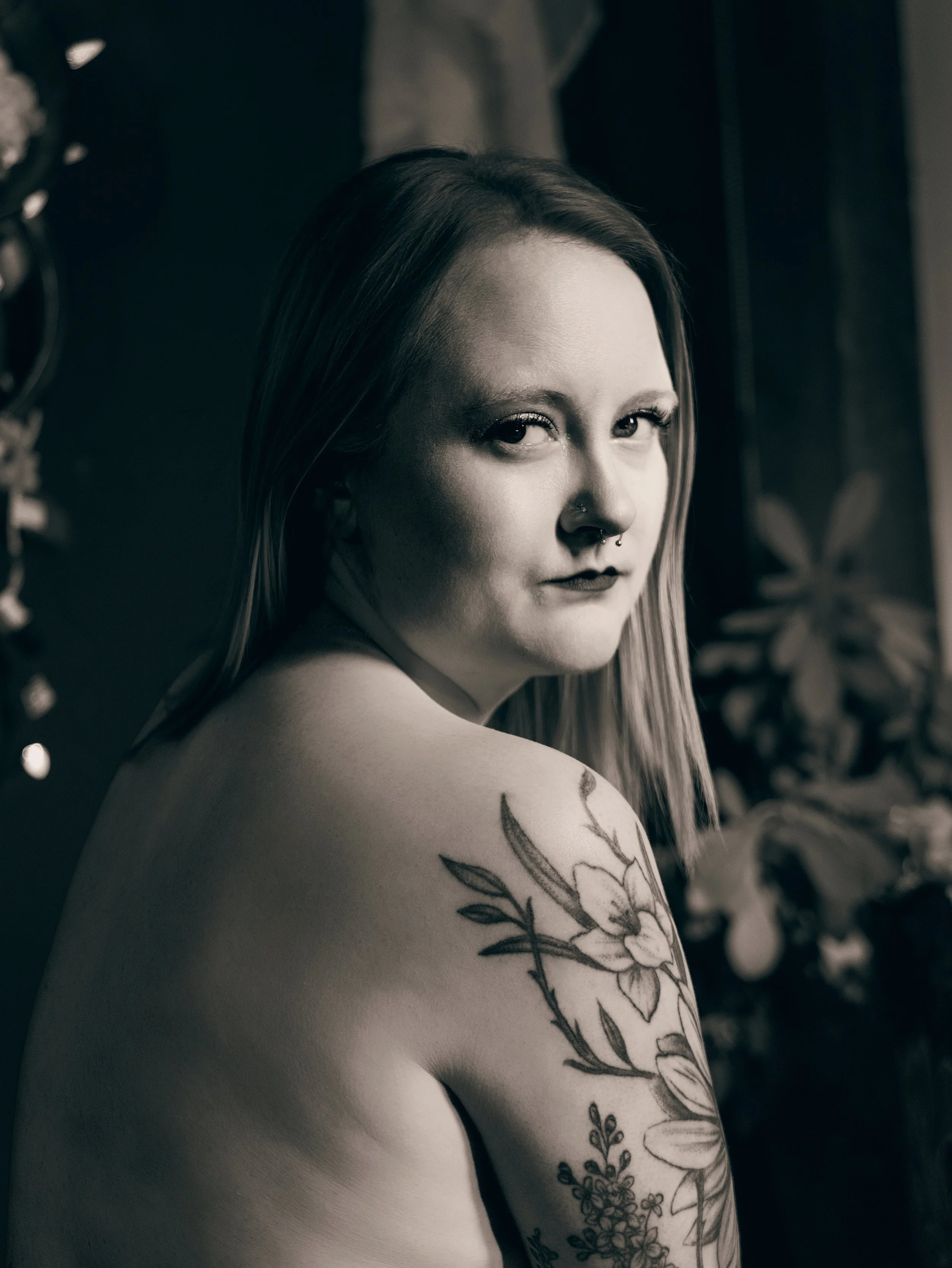 A young woman with long hair and tattoos on her shoulder and arm, looking over her shoulder at the camera, in black and white.