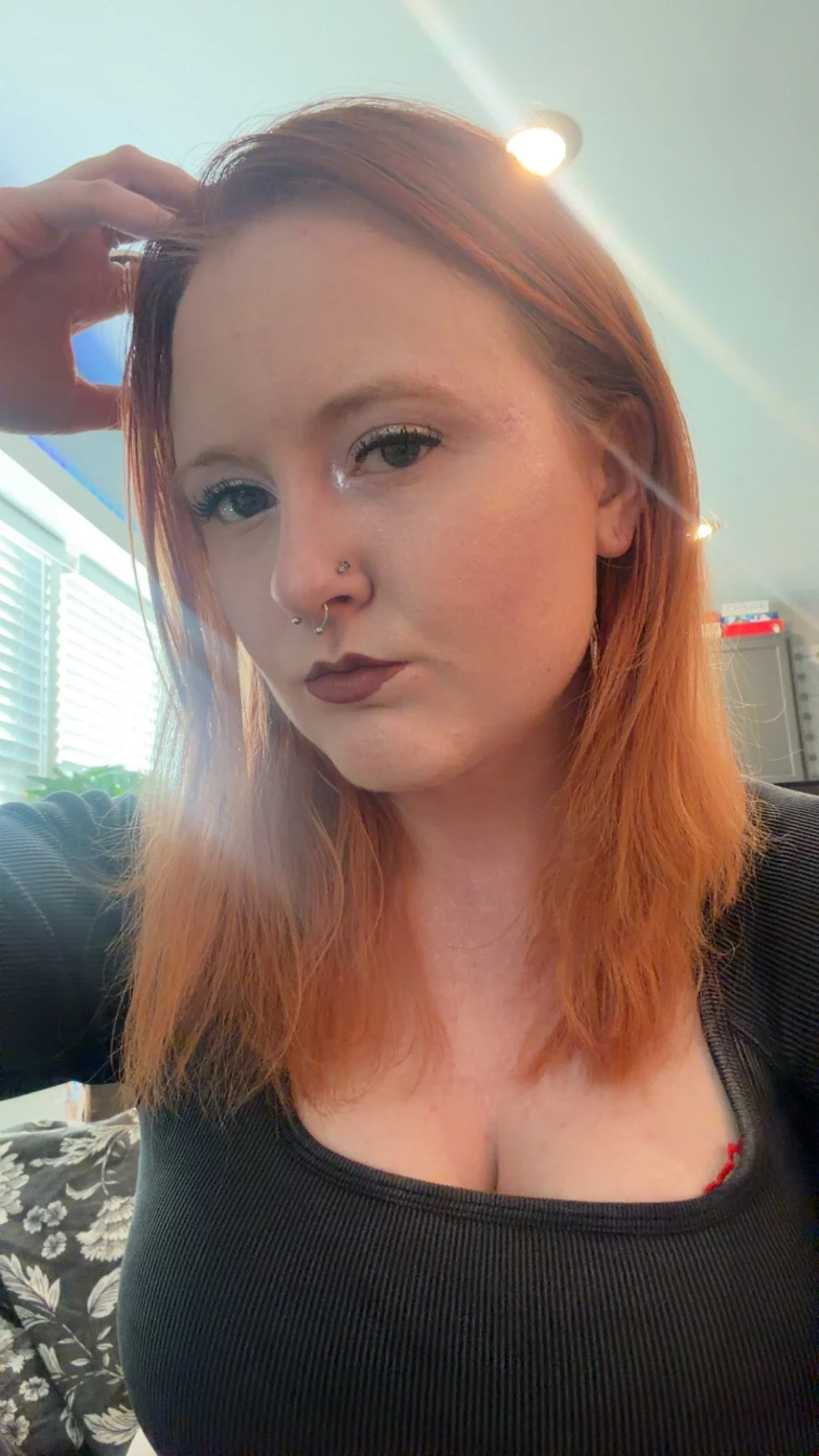 A woman with reddish hair and light skin posing indoors, wearing makeup and a black top, with a nose piercing, in front of a window with blinds and a room with shelves and boxes in the background.