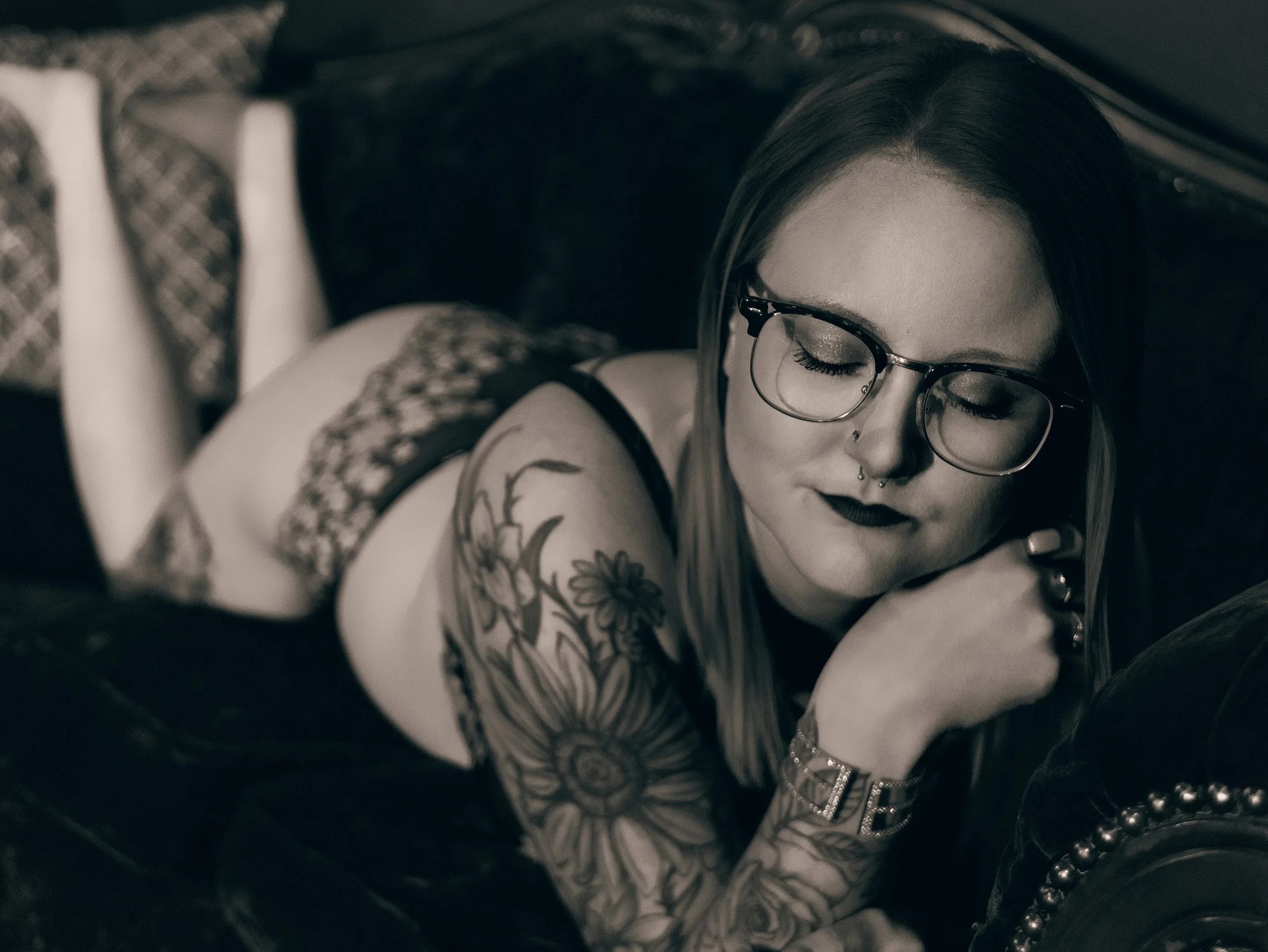 A woman with dark hair and glasses, wearing dark lipstick and makeup, lying down with her eyes closed surrounded by various necklaces and a tattoo on her arm. The photo is in black and white.