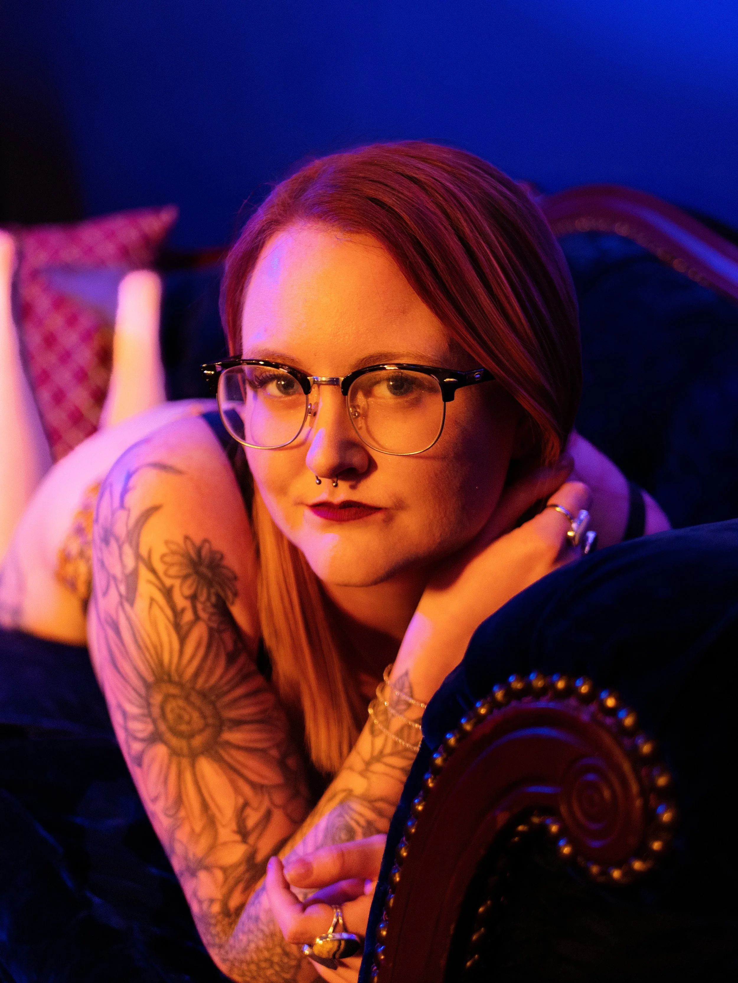 Close-up of a woman with red hair and glasses, lying on her side with her hand near her face, tattoos on her arm, and adorned with jewelry, in a dimly lit room with blue and purple lighting.