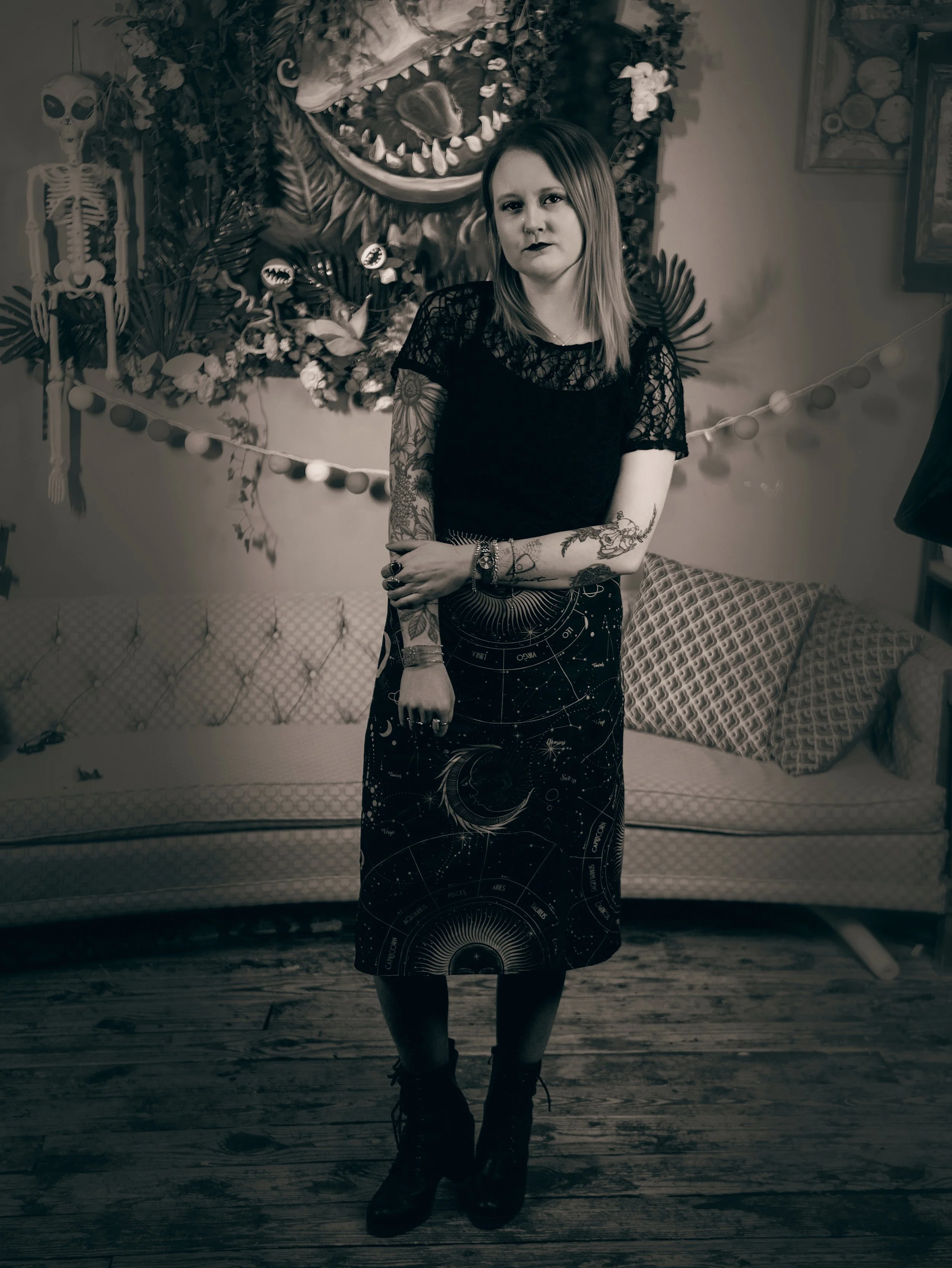 A woman with tattoos wearing dark clothing and combat boots stands in front of a decorative background with a large dinosaur head, a skeleton, floral arrangements, and hanging lights in black and white.