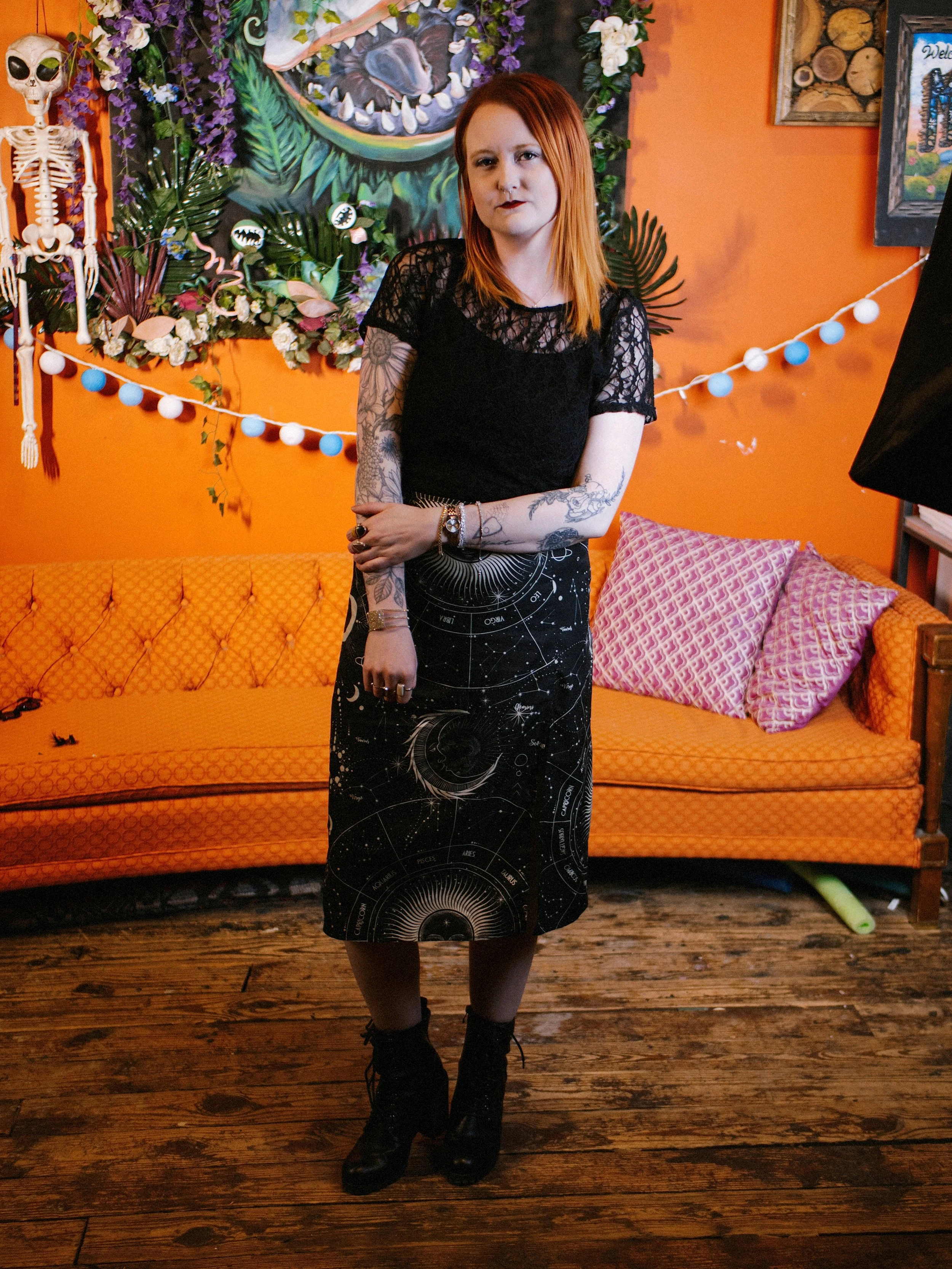 A woman with red hair and tattoos on her arms is standing in front of a colorful, artistic background with a large, painted animal mouth and greenery. She is dressed in a black dress with a celestial and zodiac design, and is wearing black combat boo