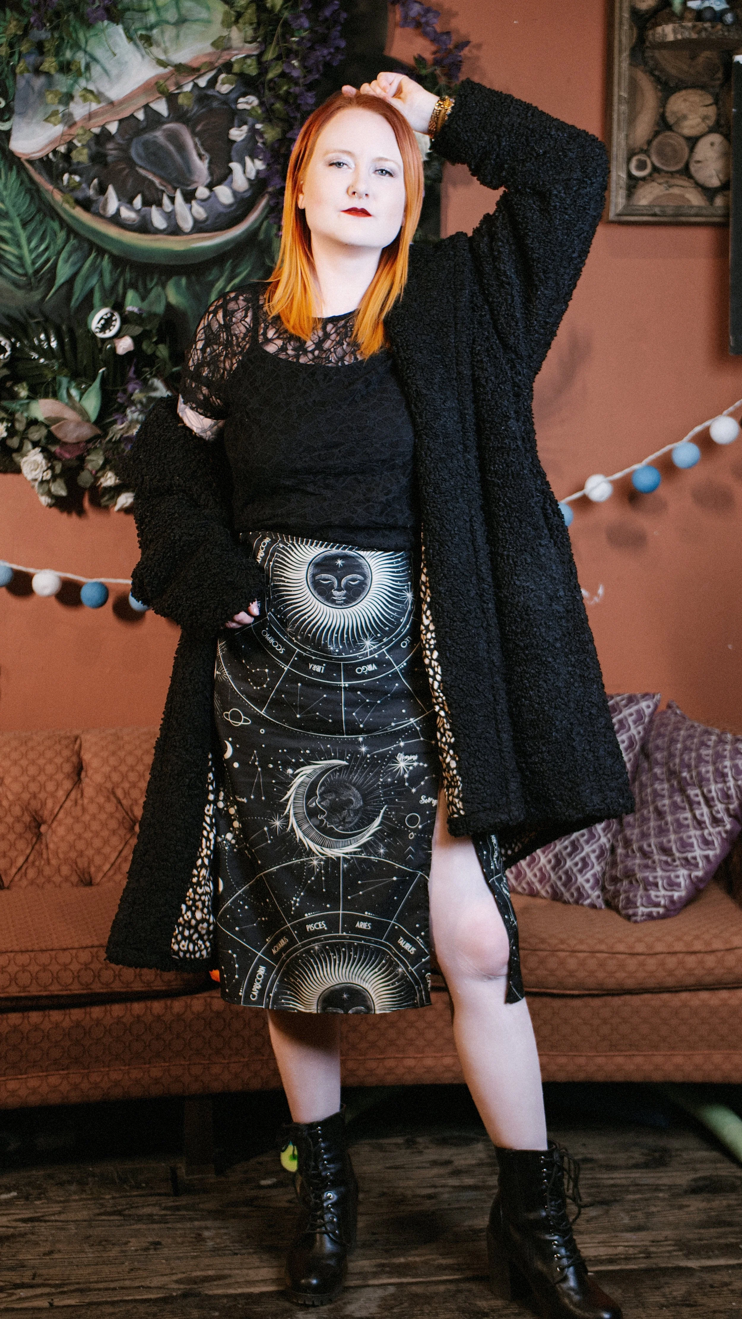 A woman with red hair and fair skin standing in a cozy interior, wearing a black lace top, a celestial-themed skirt with zodiac signs, a textured black coat, and black combat boots. She has one hand on her head and the other in her pocket, with a con
