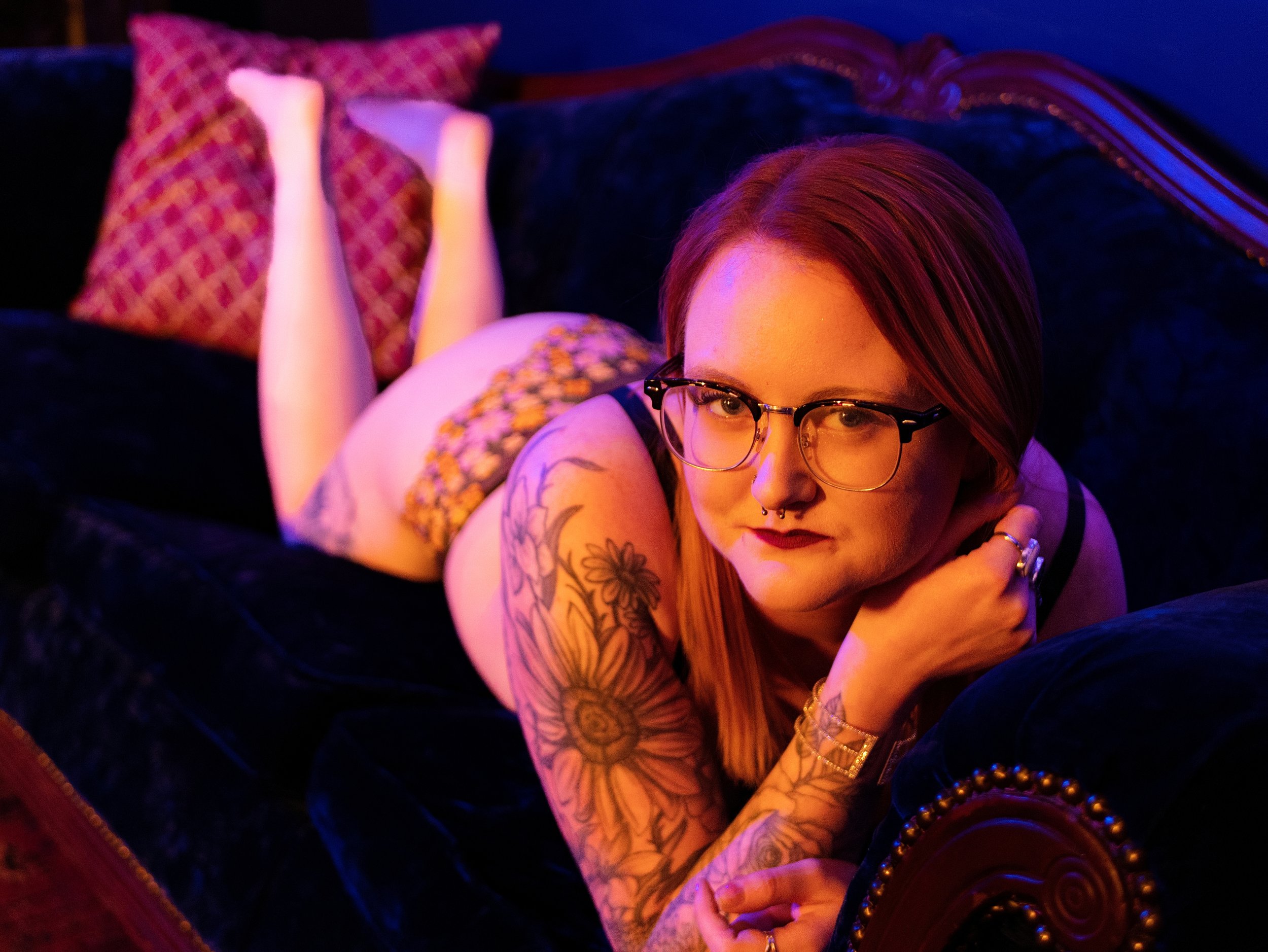 A woman with red hair, glasses, and tattoos lying on her stomach on a dark couch in a colorful, dimly lit room with pink and purple lighting, wearing a floral top and black pants.