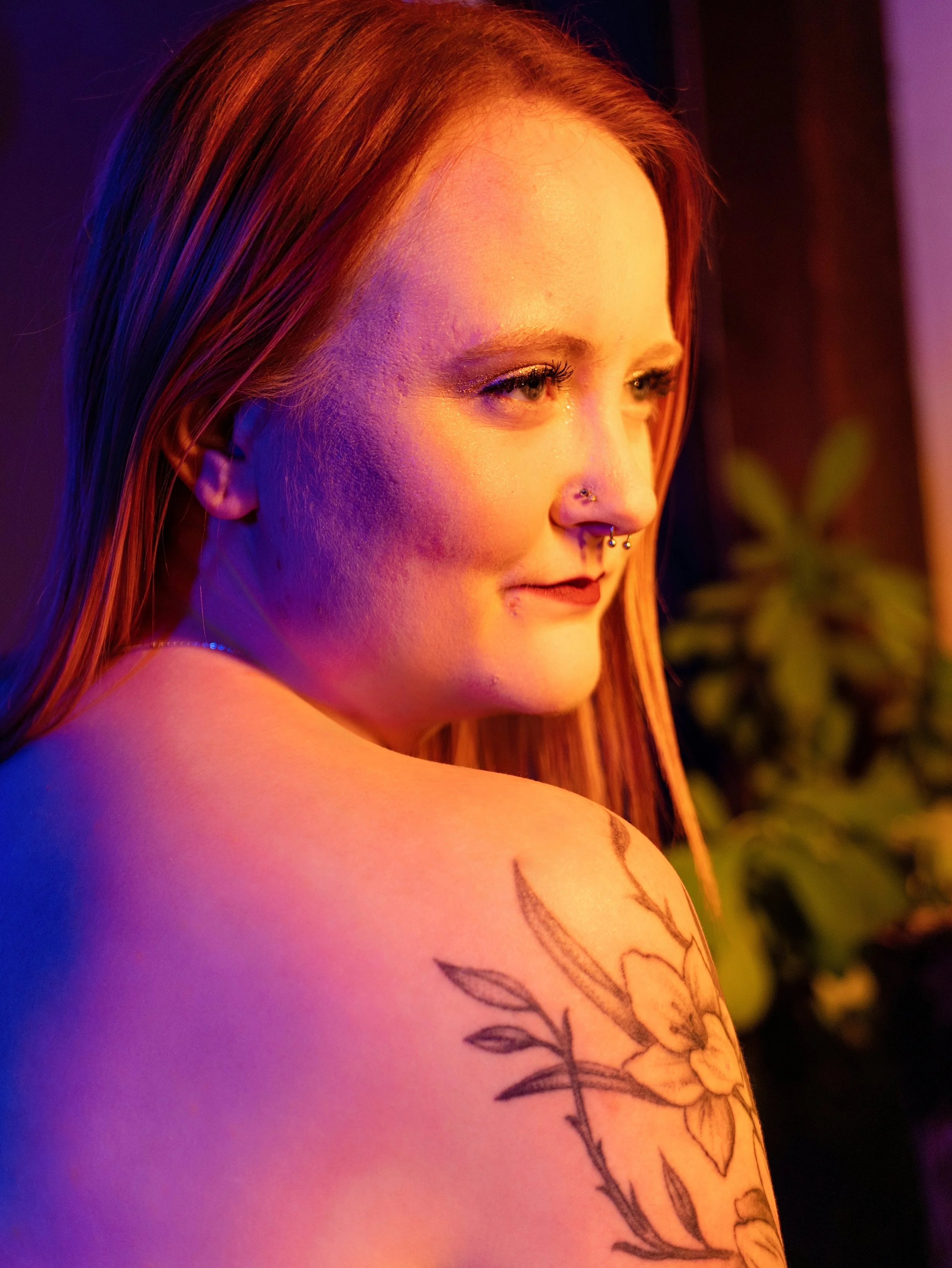 Close-up of a woman with light skin and red hair, showing her face and shoulder with a tattoo of flowers and leaves. She has piercings on her nose and lip and is illuminated by warm and purple colored lighting.
