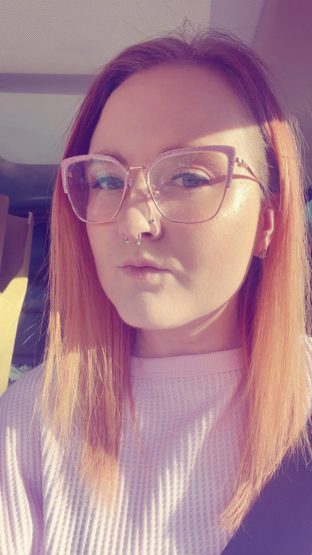 A woman with long orange hair wearing large pink glasses, a septum piercing, and earrings, looking at the camera inside a sunlit room.