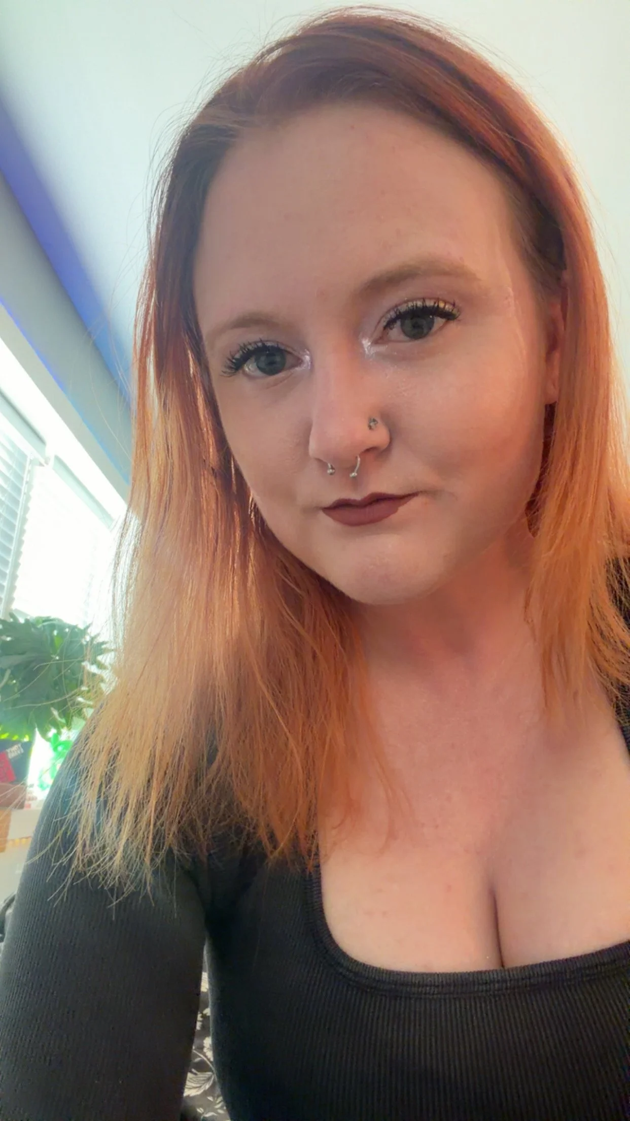 A woman with reddish hair and piercings, wearing a black top, taking a selfie indoors near a window with blinds and some green plants visible in the background.