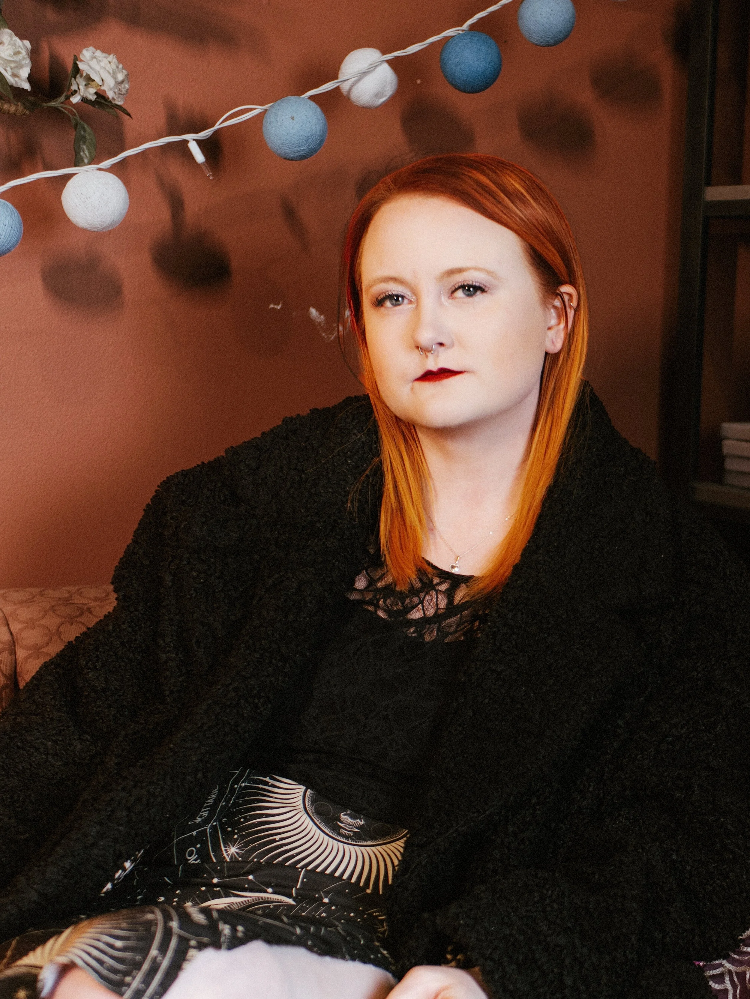 A woman with red hair, pale skin, and makeup including dark lipstick and eyeliner, wearing a black fuzzy sweater and a necklace, is sitting in front of a brick red wall decorated with a string of white and blue globe string lights.