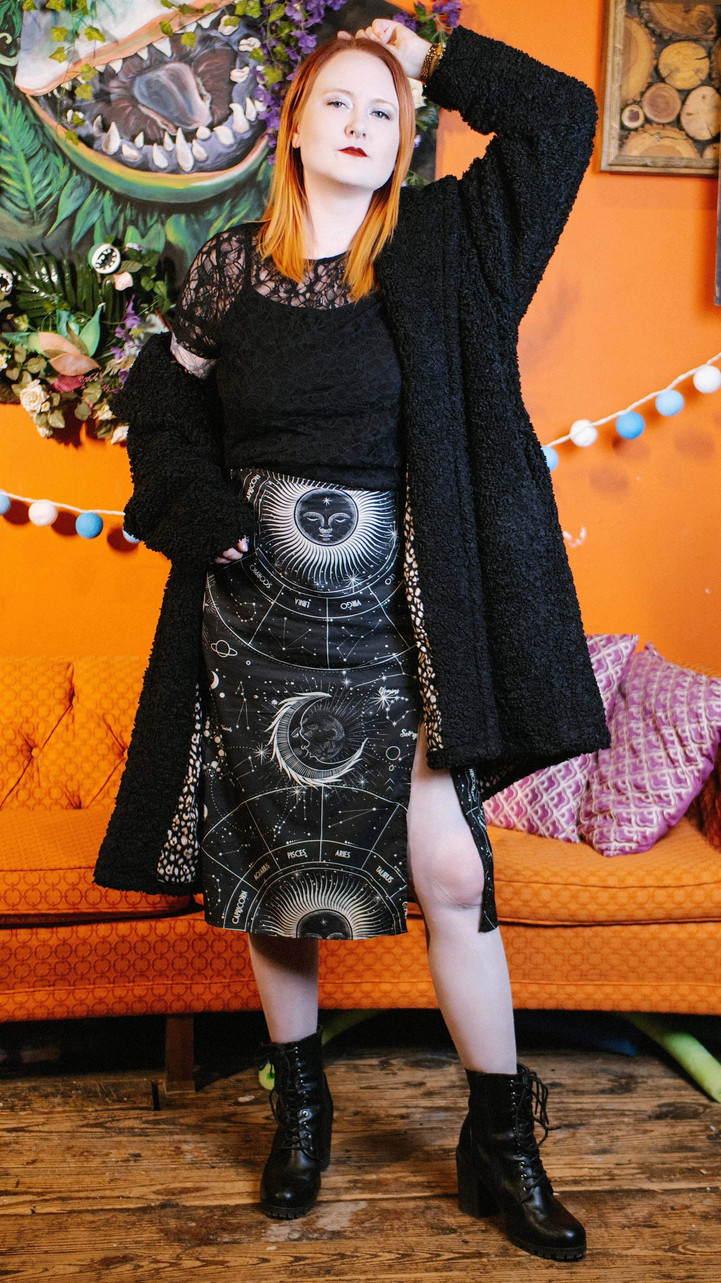 A woman with red hair and fair skin standing indoors in front of an orange wall adorned with artwork and string lights. She is wearing a black lace top, a long skirt with celestial and zodiac designs, a long black coat, and black lace-up boots. She h