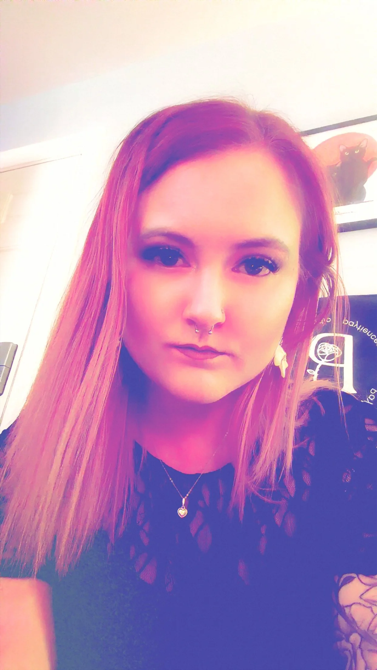 A young woman with long reddish hair, septum piercing, wearing a black lace top and a heart-shaped necklace. There is a framed picture of a black cat on the wall behind her.