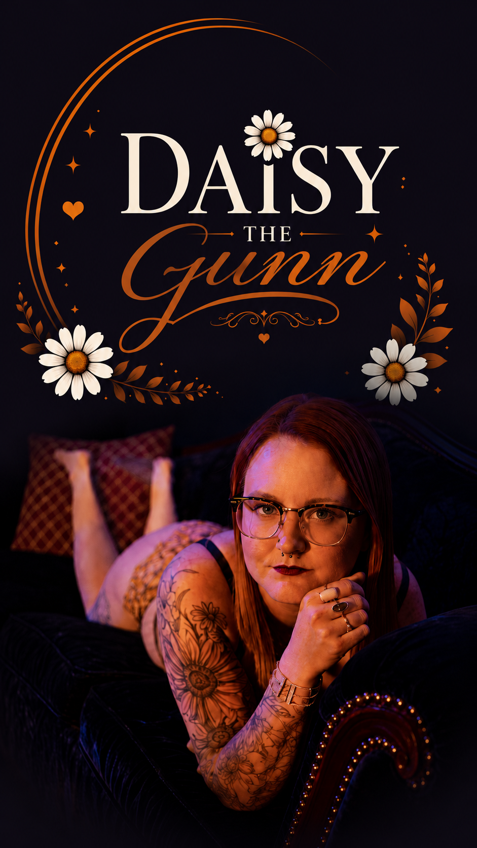 A woman with tattoos, glasses, and dark lipstick lying on a black velvet couch with a dark background, with a book titled "Daisy the Gumn" decorated with flowers and hearts hanging above her.