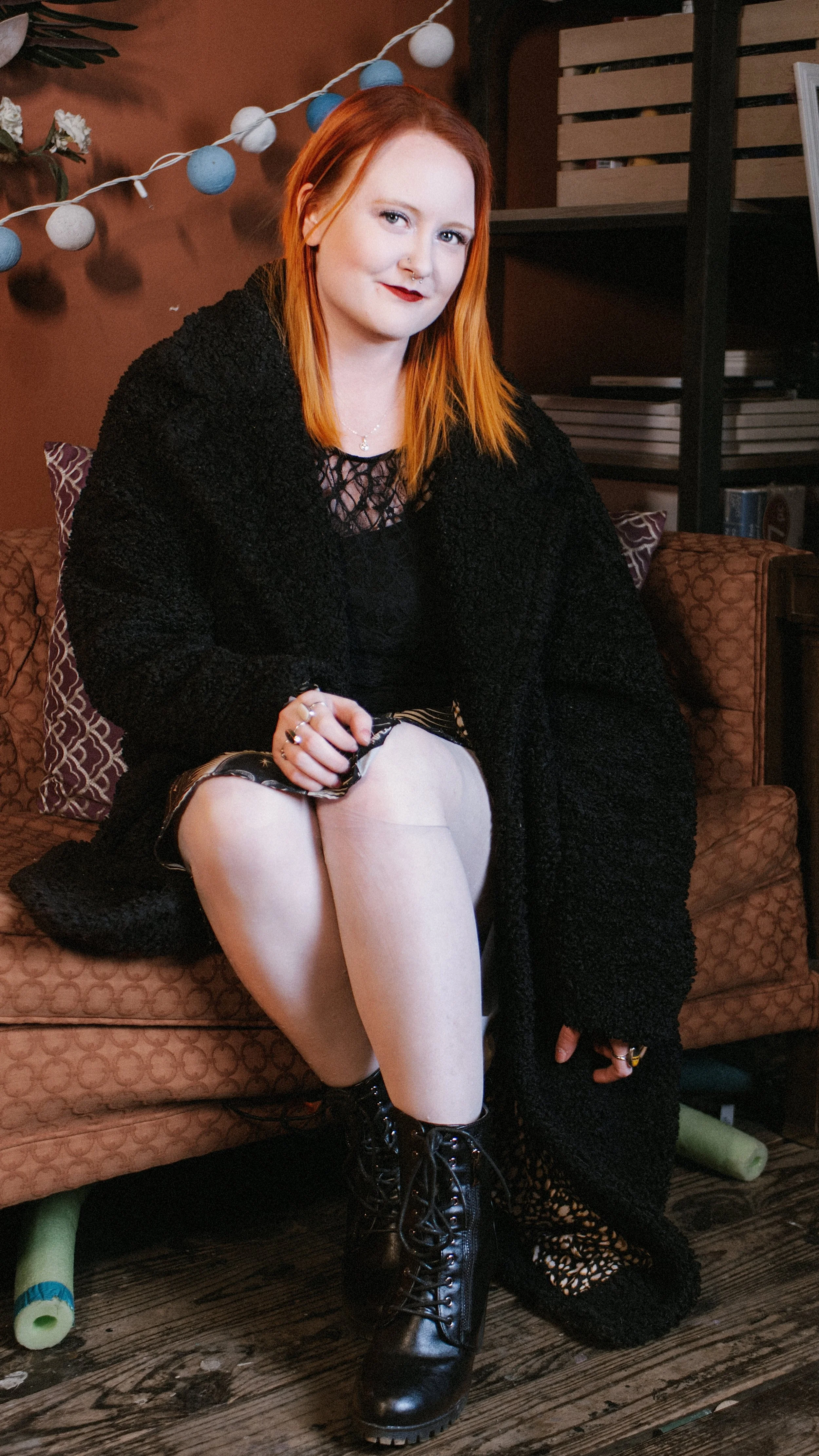 A young woman with red hair sitting on a patterned sofa with her legs crossed, wearing a black jacket, a black lace top, white tights, and black lace-up combat boots. She has a septum piercing, dark lipstick, and jewelry. There are string lights and 