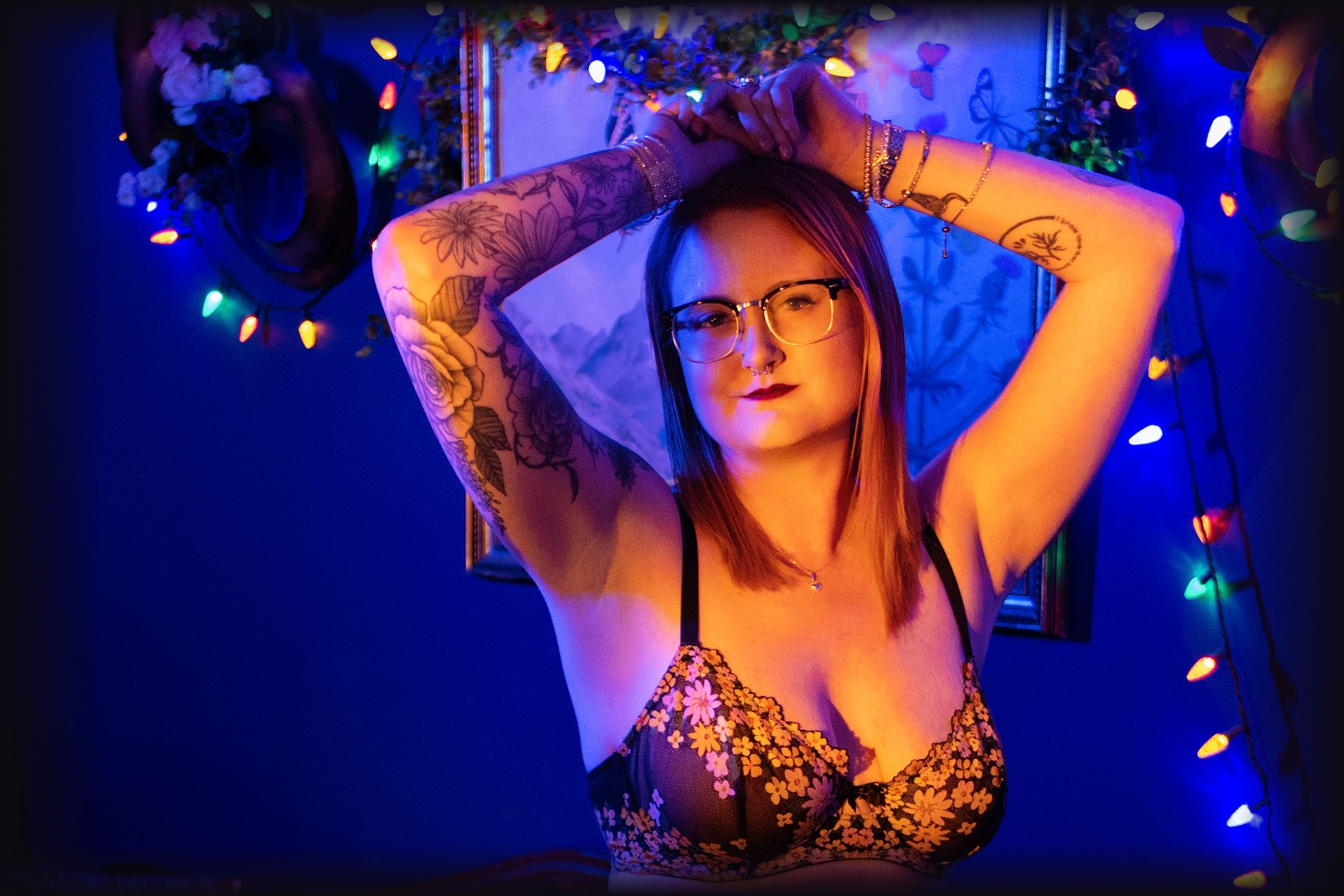 A woman with glasses and tattoos on her arms is posing with her hands behind her head in a room decorated with colorful string lights and a wall hanging featuring a moon and butterflies.