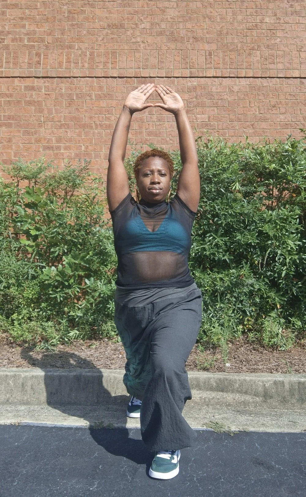 Woman practicing yoga outdoors against a brick wall and green bushes, wearing black and blue athletic clothing with her arms raised above her head and one leg bent.