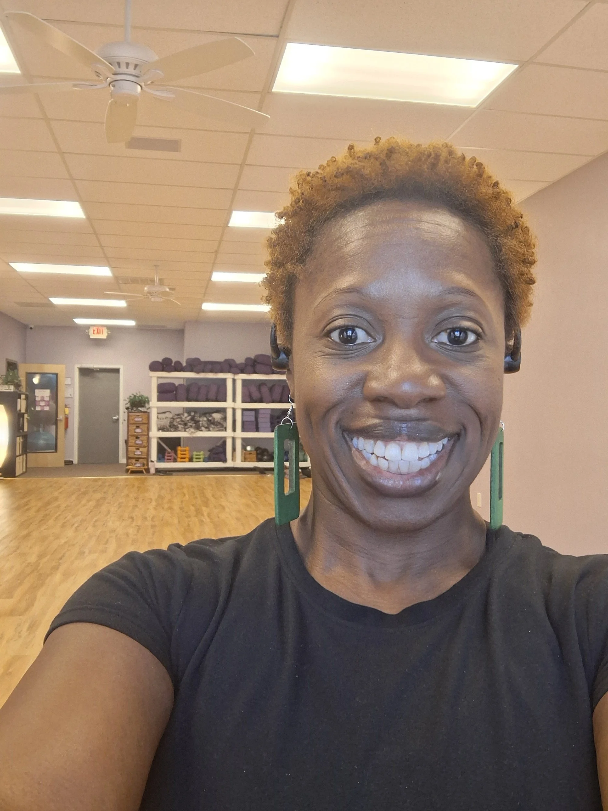 A woman in a black shirt smiling in a gym studio with purple exercise mats and shelves in the background.