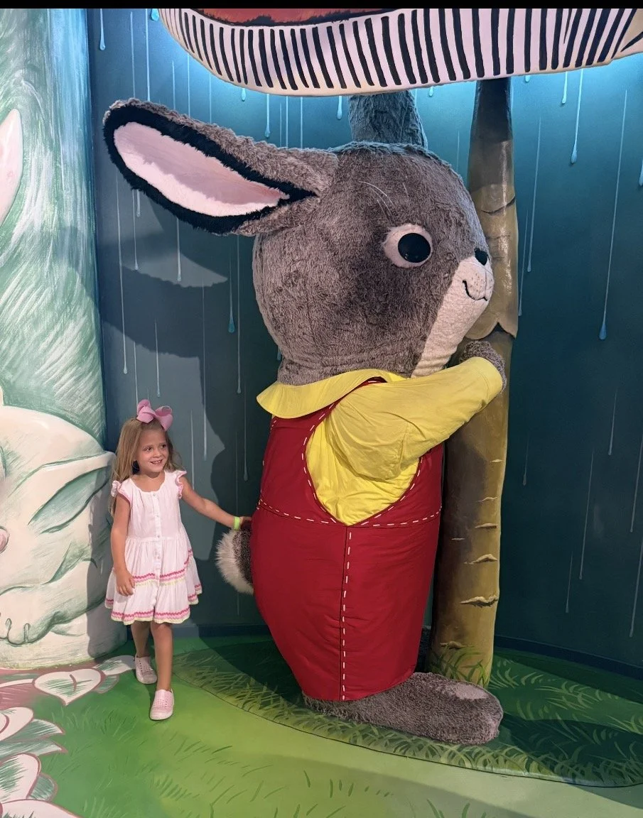A young girl with a pink bow in her hair standing next to a large, plush rabbit mascot dressed in a red and yellow outfit, hugging a tree in a colorful, cartoon-themed indoor setting.