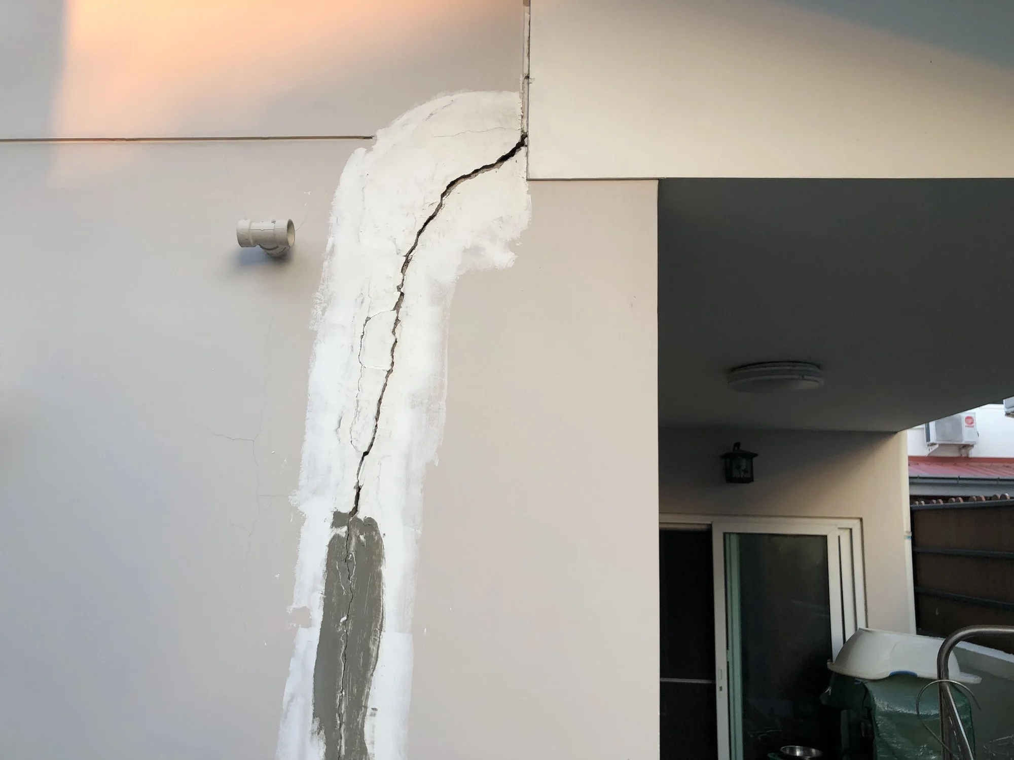 Image of a wall with visible surface cracks and structural damage, along with a small pipe protruding from the wall, indicating potential issues with the building's foundation or structure.