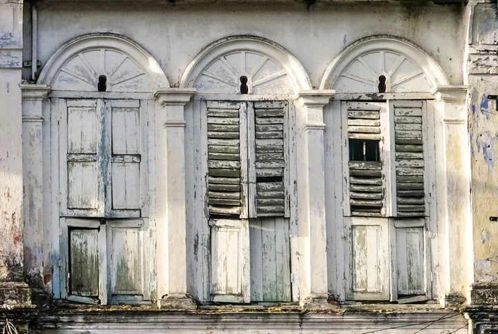 Three sets of old, weathered wooden window shutters, with peeling paint, on a building's facade.