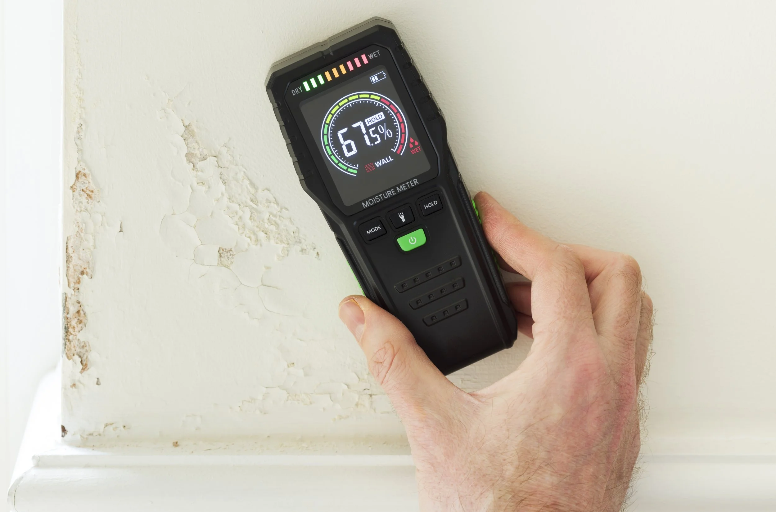 A moisture meter showing a reading of 67.5% in front of peeling paint or drywall on a wall.