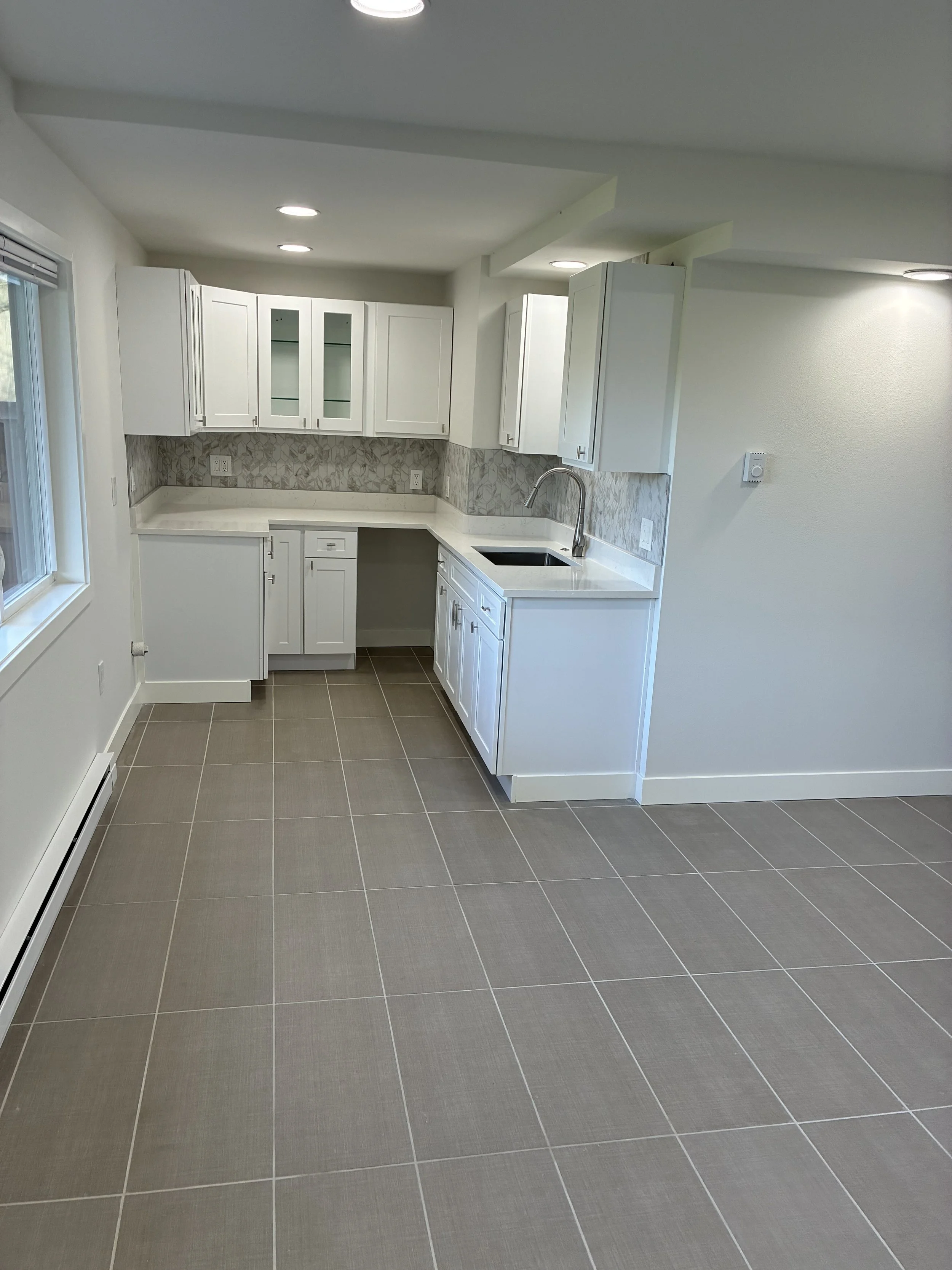 Empty modern kitchen with white cabinets, beige and gray patterned backsplash, stainless steel sink, and gray tiled floor.