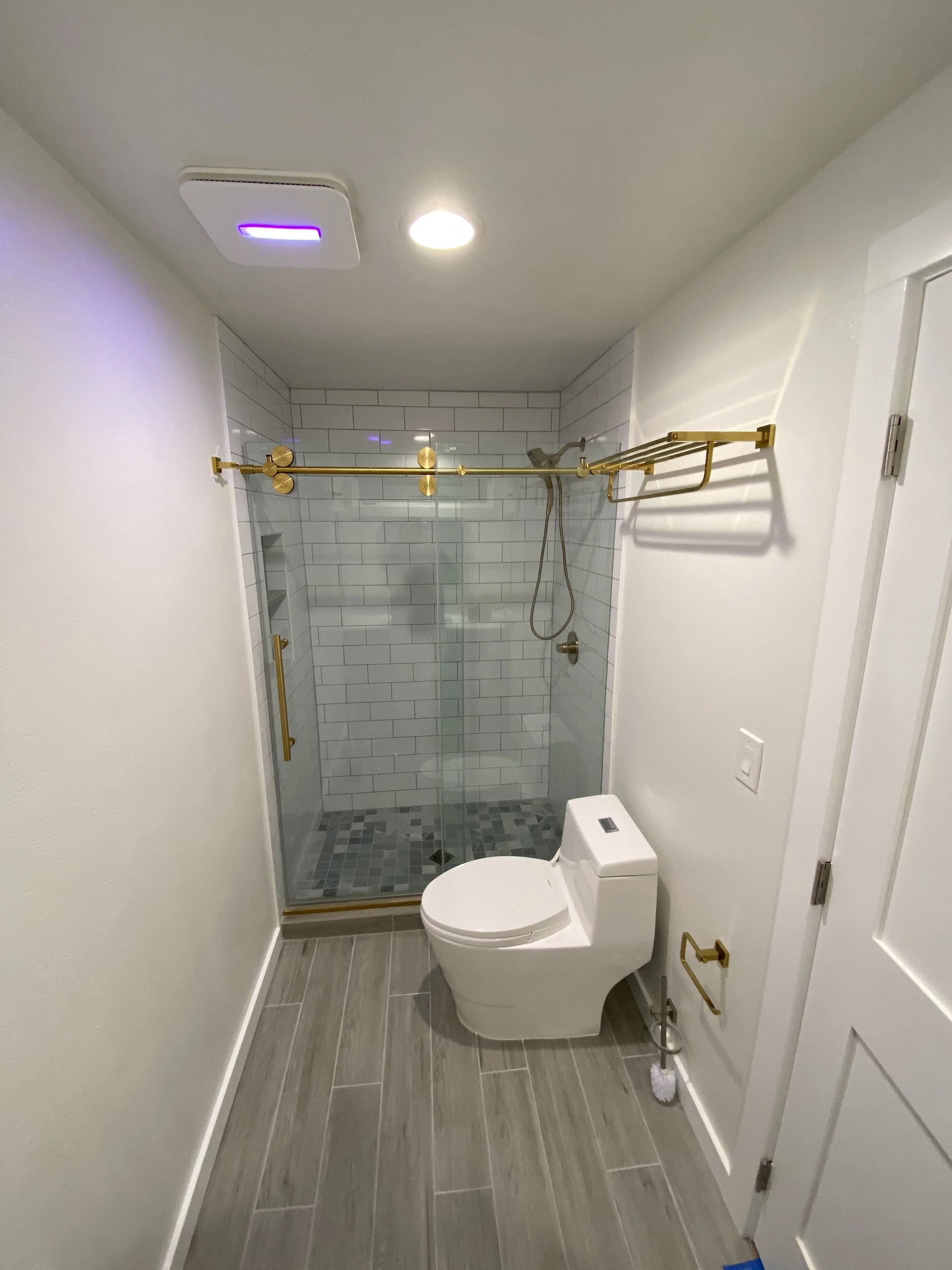 A modern bathroom with a shower enclosure with a glass door, white subway tile walls, gold fixtures, a mounted shower head, and a toilet. The floor has gray tiles, and there is a white wall with a gold towel rack and a white door.