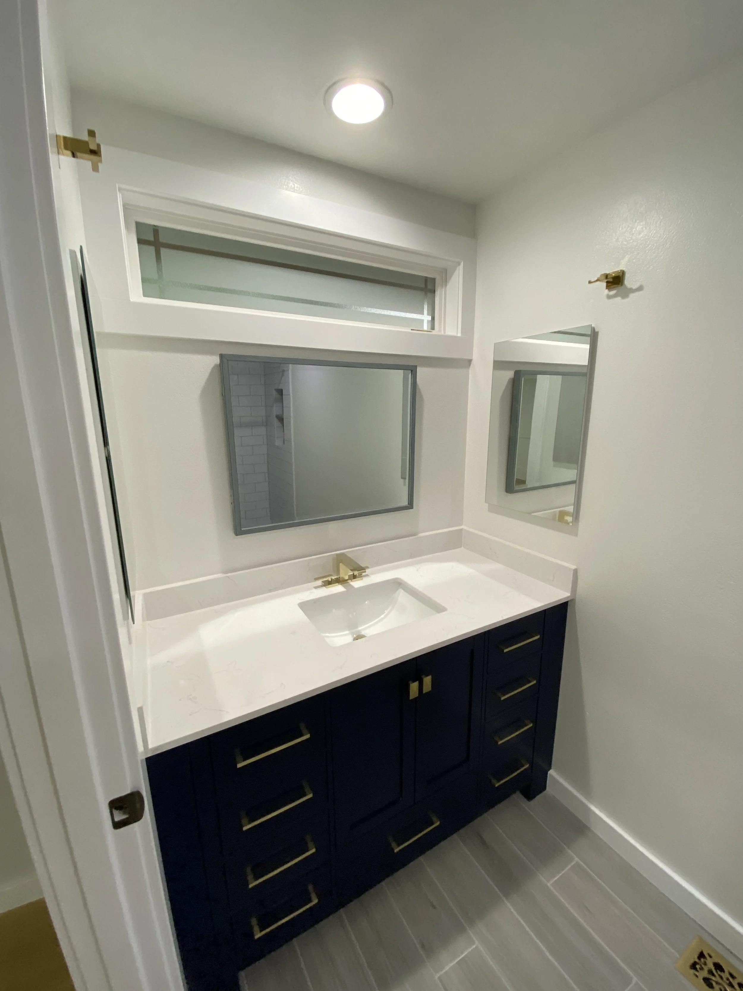 A modern bathroom vanity with navy blue cabinets, gold hardware, a white marble countertop, a rectangular undermount sink, two mirrors, and gold fixtures. There is a small window and a light fixture on the ceiling.