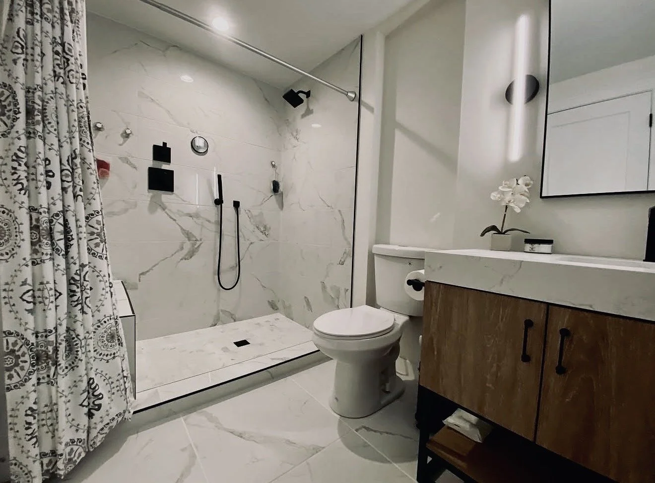 Modern bathroom with a walk-in shower featuring a marble wall and a shower head, a toilet, and a vanity with a mirror and decorative flower. The flooring and wall tiles are white with gray veining.