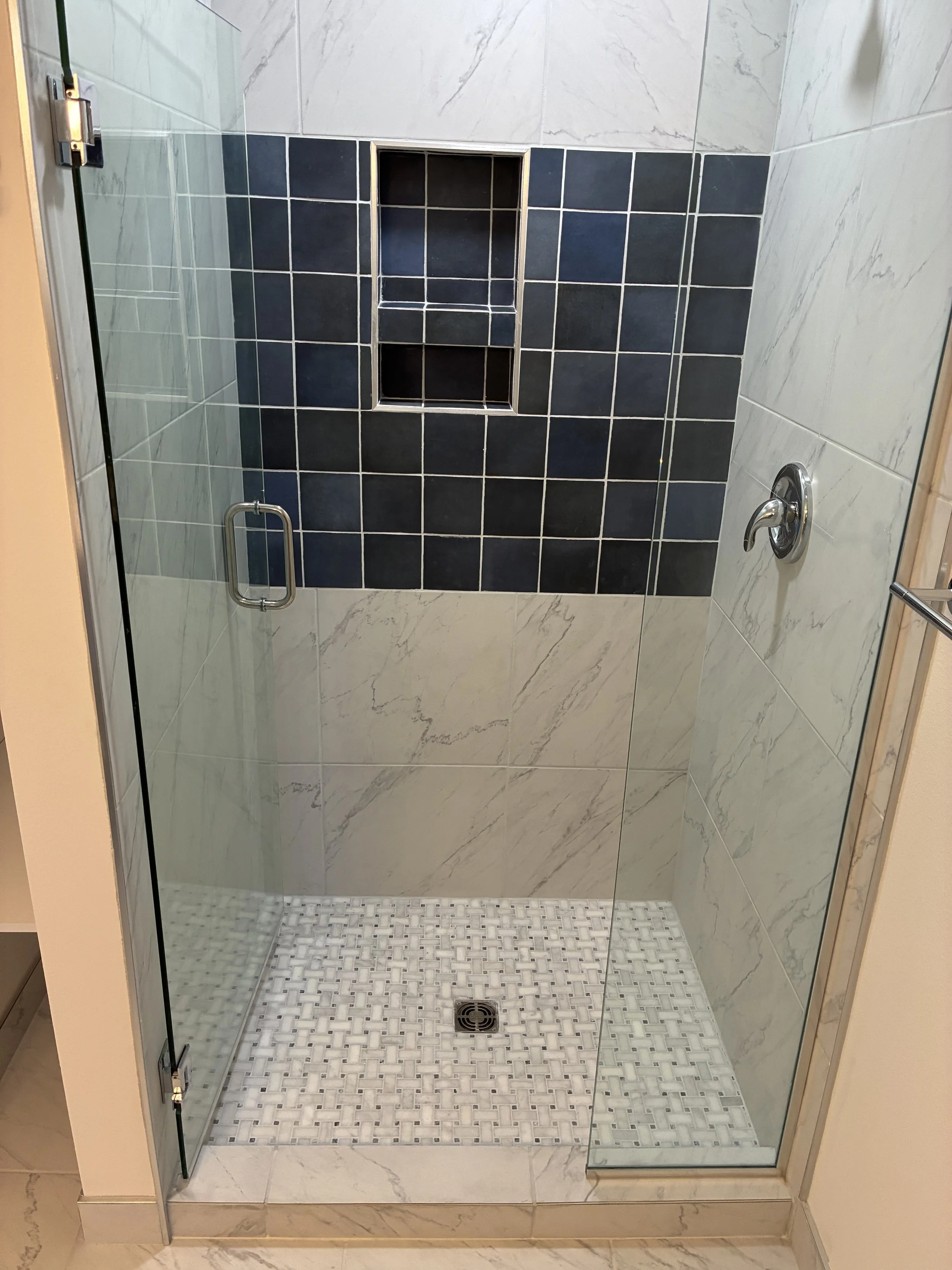 A modern glass shower stall with a black tiled accent wall inside. The shower has a silver handle on the door and a silver shower faucet on the right wall. The shower floor has small white and gray tiles with a drain at the center. There's a built-in niche in the black tiled wall for storage.
